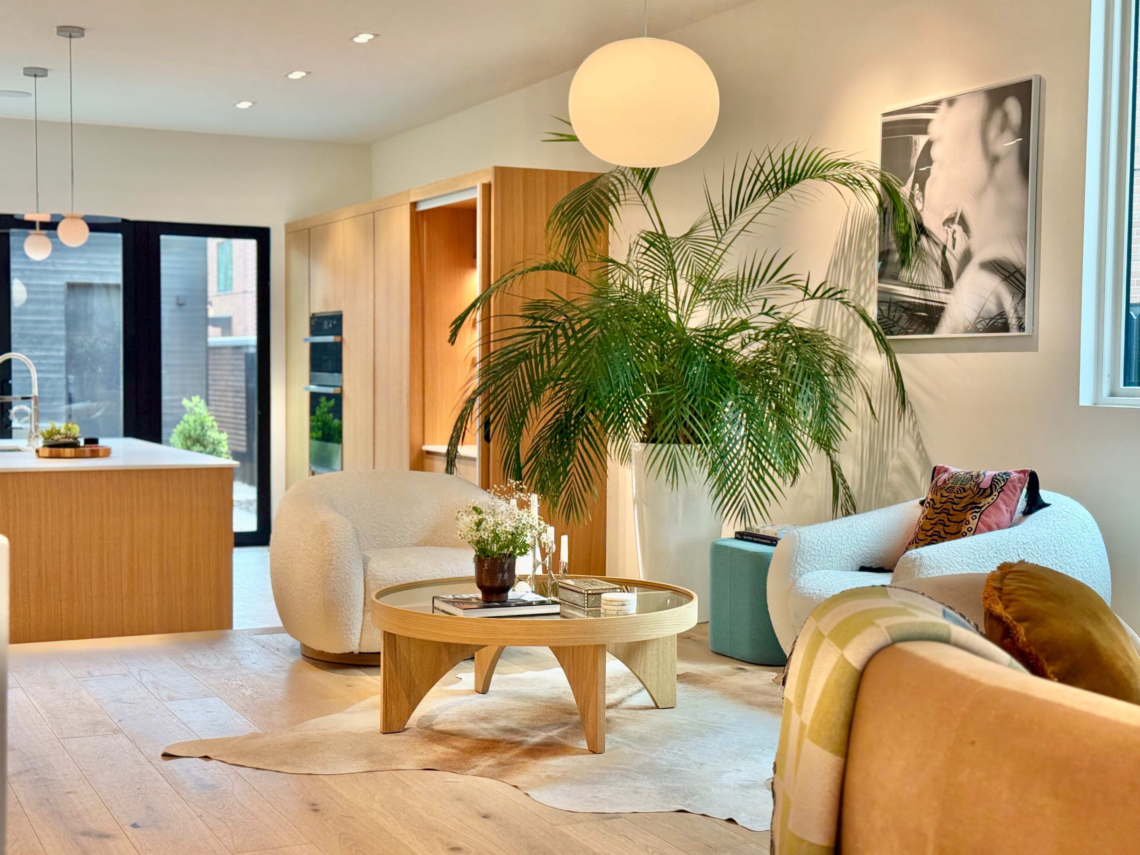 The image shows a modern living room with a circular coffee table, potted palm, and plush seating arranged around a light-colored area rug.