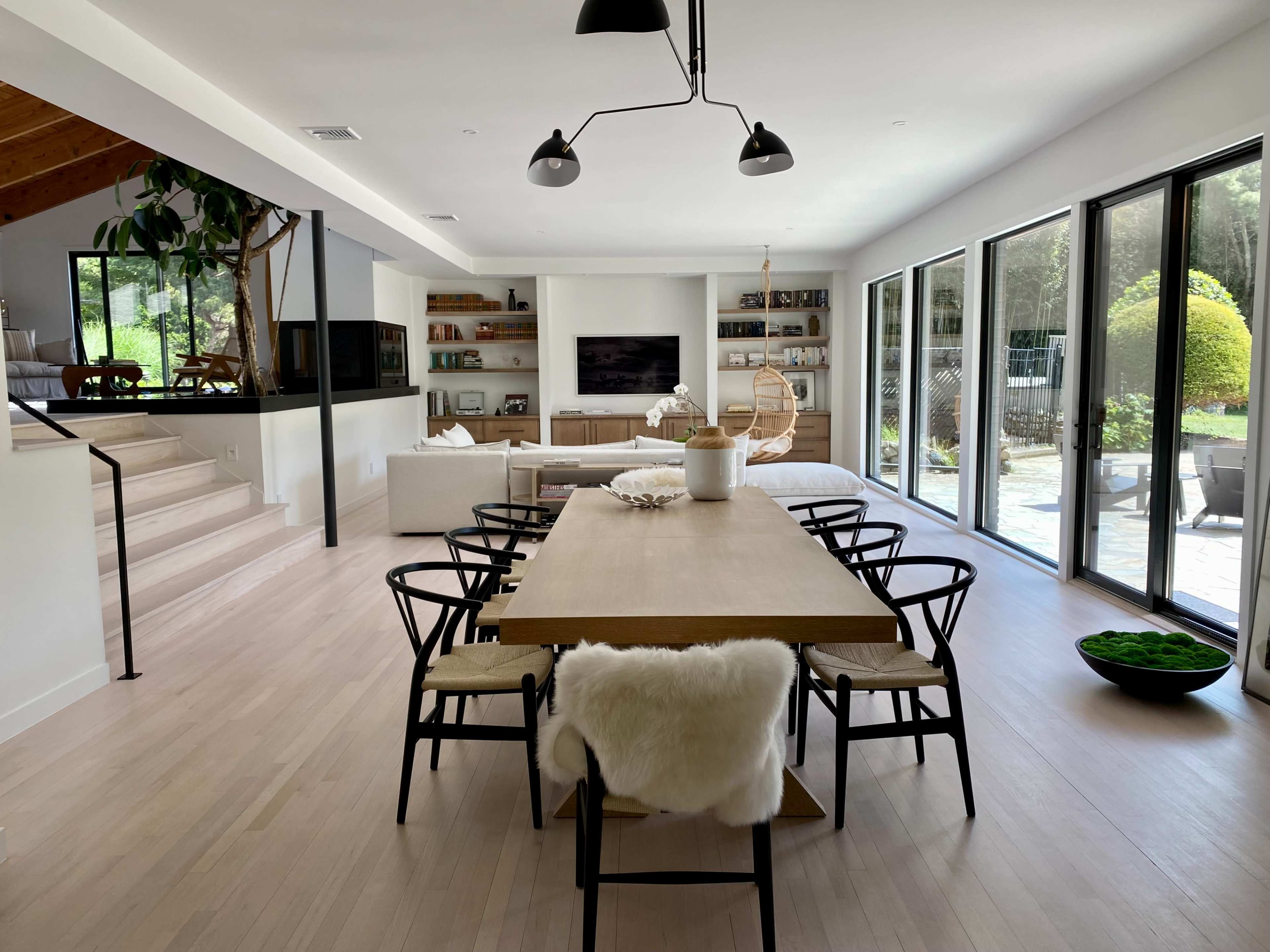 The image shows a spacious, modern dining area with a long wooden table surrounded by black chairs, leading to large glass doors that open to an outdoor patio.
