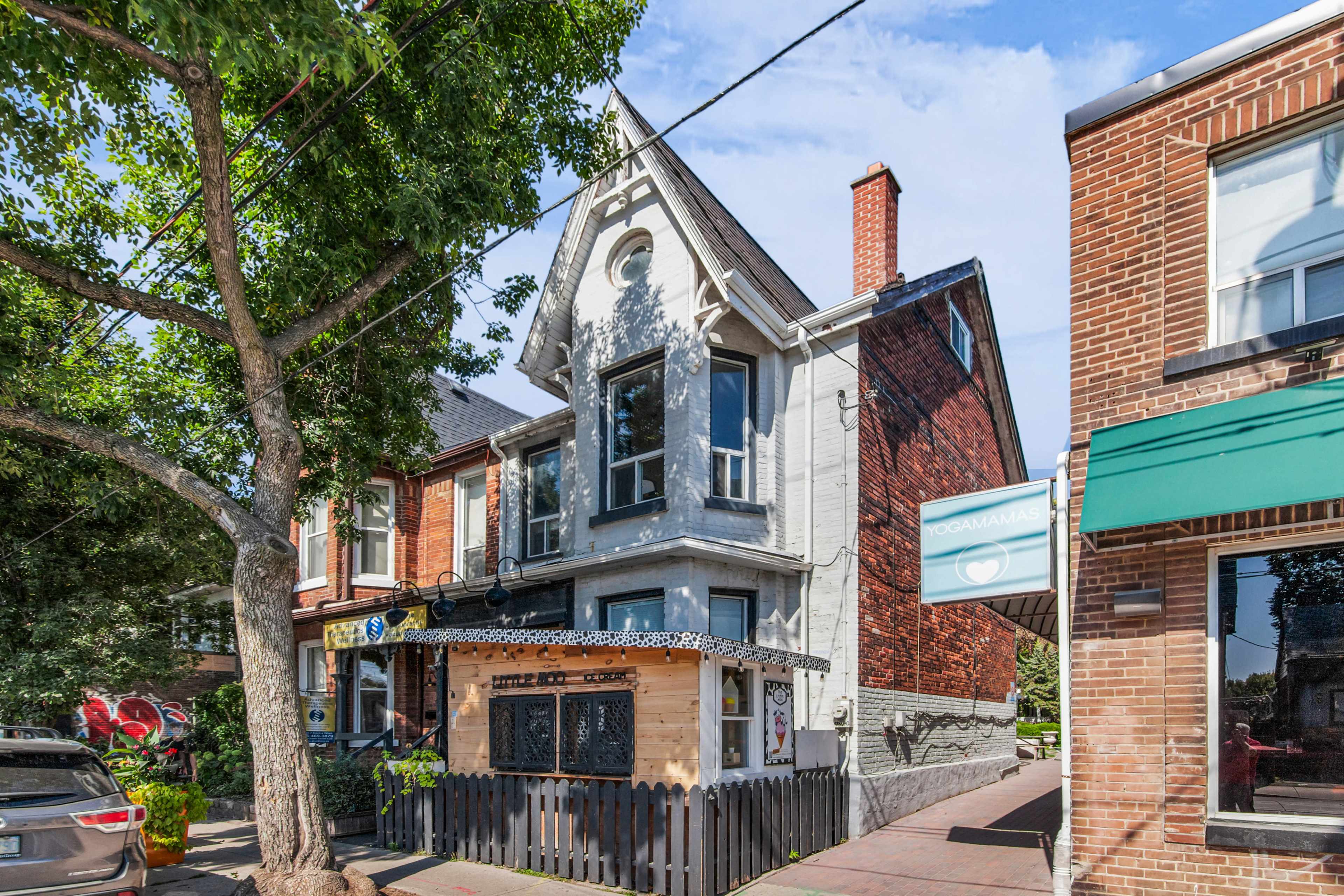 The image shows a two-story building with a peaked roof, featuring a mix of brick and gray siding, situated at the intersection of a street lined with trees and nearby commercial storefronts.