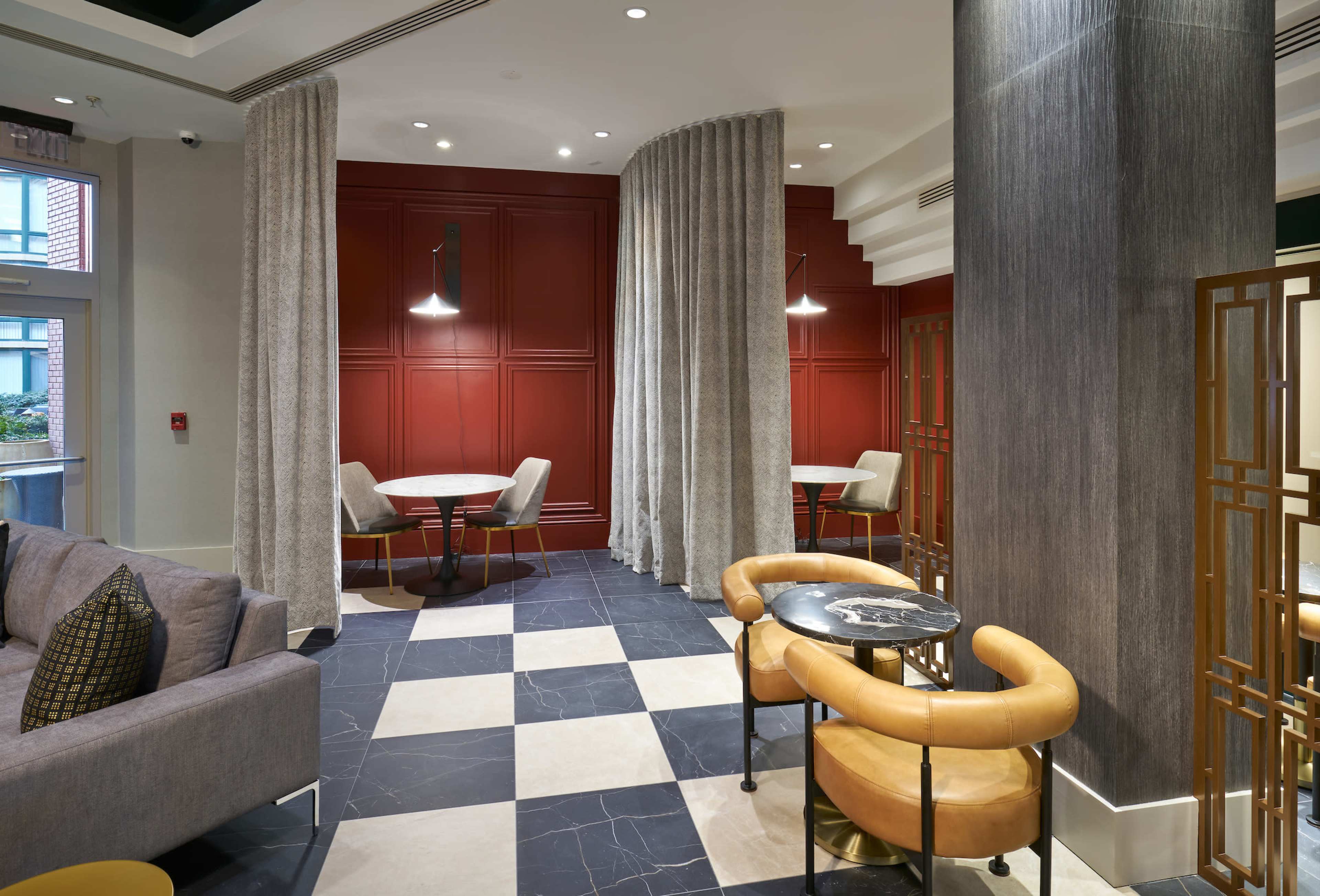 The image depicts a modern lounge area with a checkerboard floor, round tables, and upholstered seating arranged around a backdrop of red paneled walls and draped curtains.