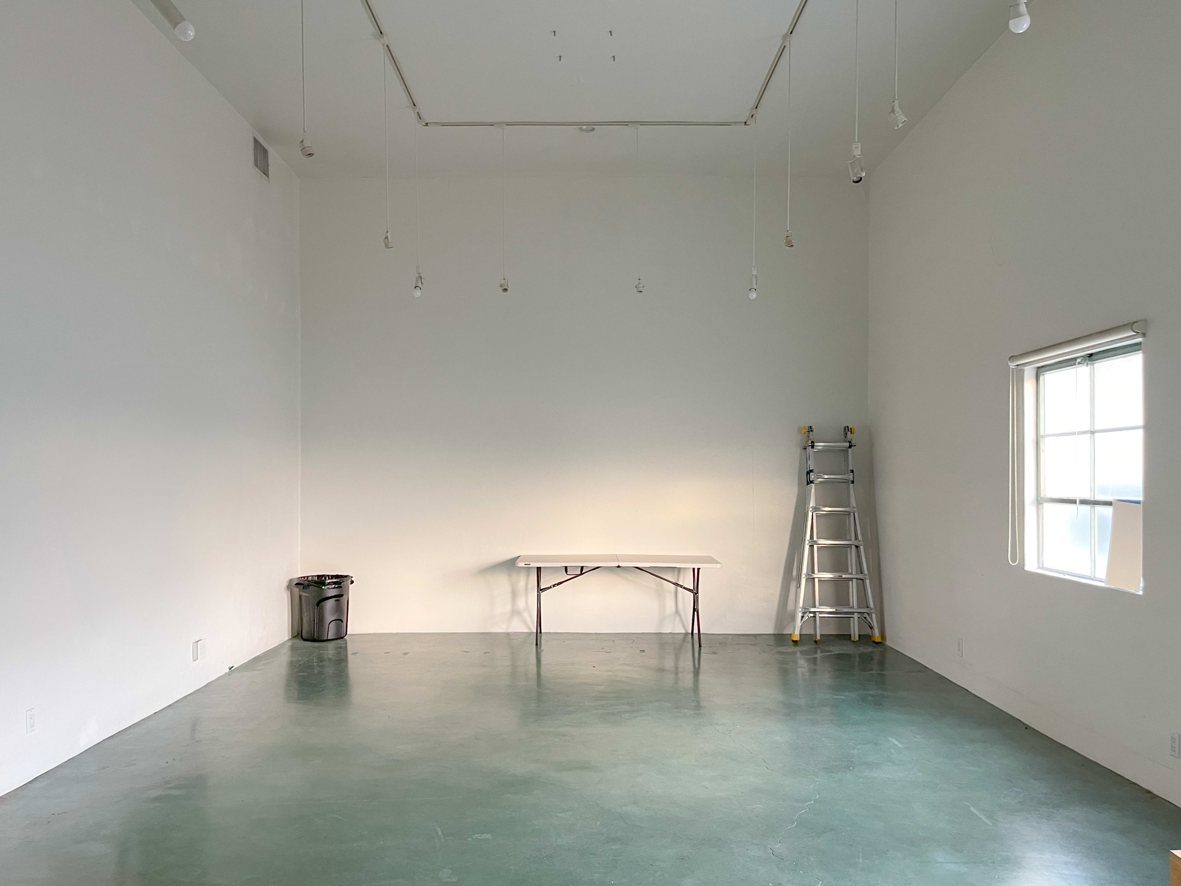 A large, empty room with a green concrete floor, a folding table in the center, a ladder against the wall, and a window on the right.