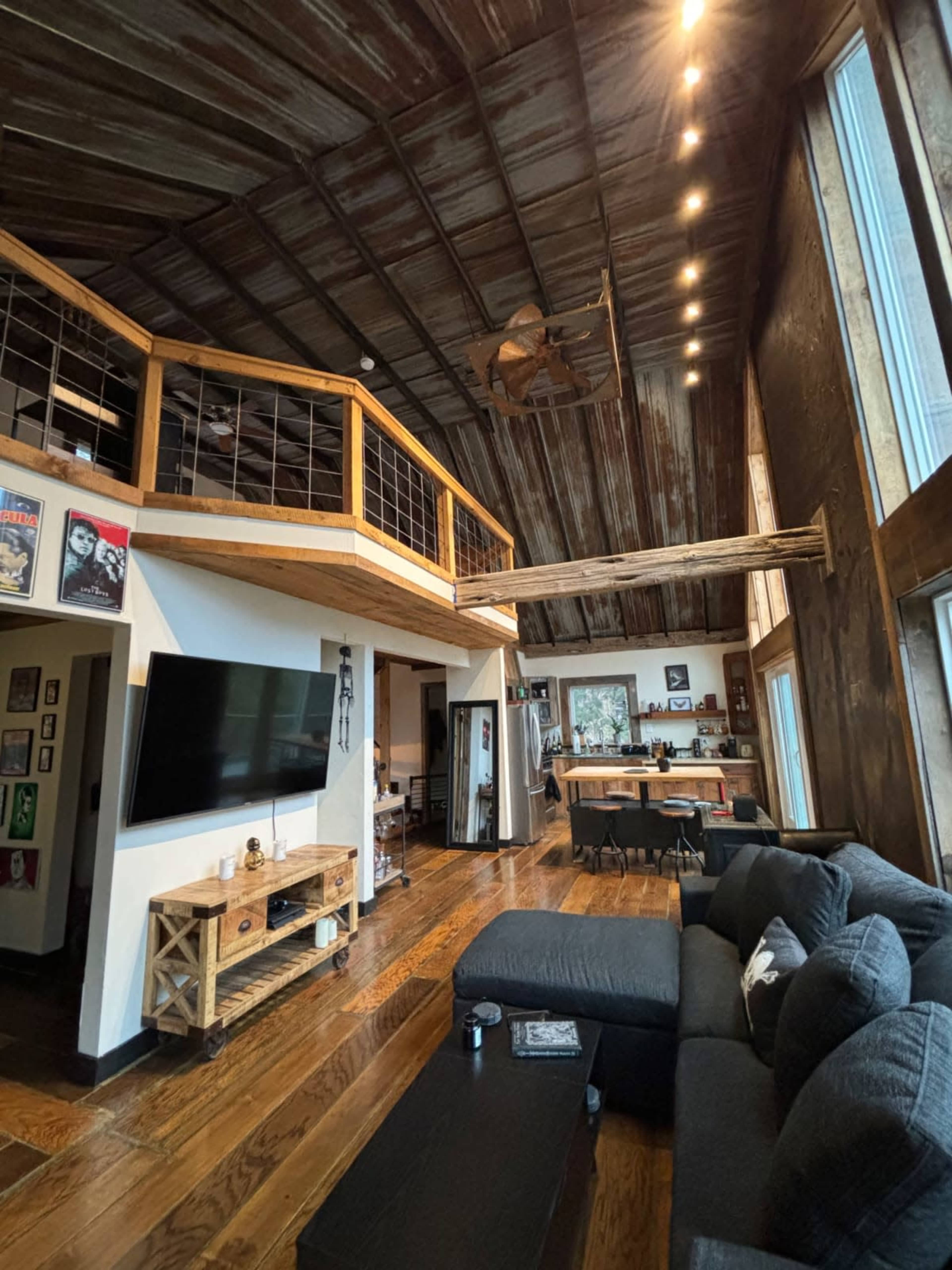 The image shows a rustic living room with a high, slanted ceiling, a wooden loft area, a dark sectional sofa, and a television mounted on a wall with framed artwork surrounding it.