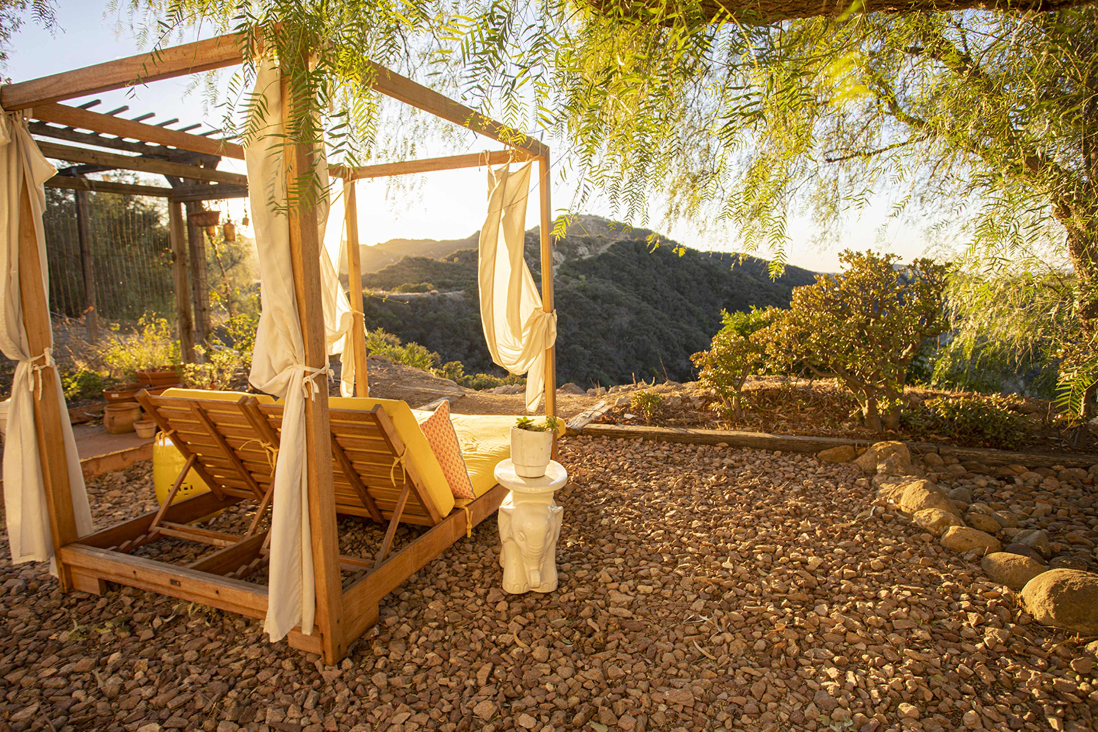 A wooden lounge area with draped fabric sits on a rocky surface, overlooking a mountainous landscape at sunset.