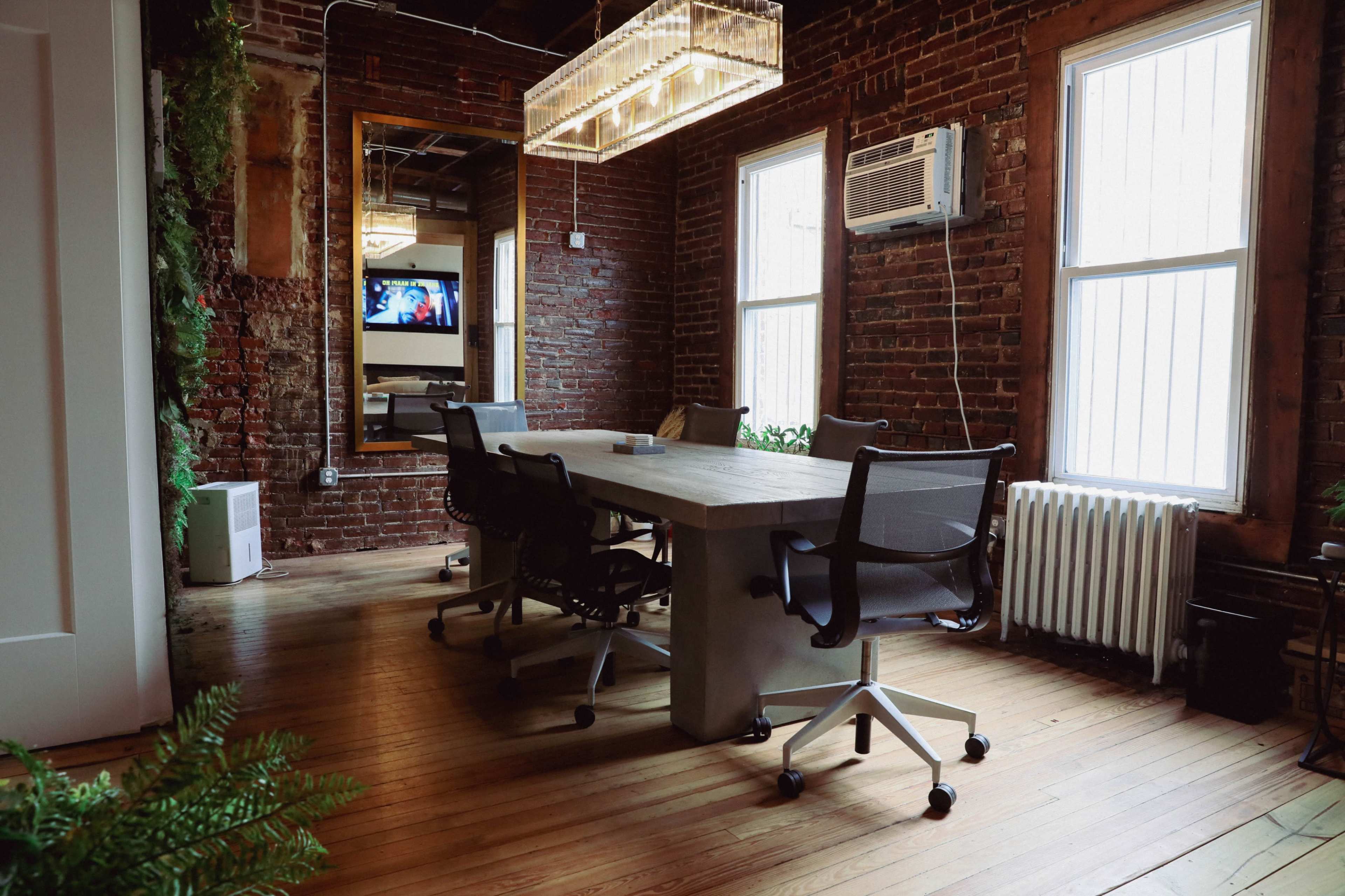 A conference room with a wooden table and several black office chairs, featuring exposed brick walls and large windows.