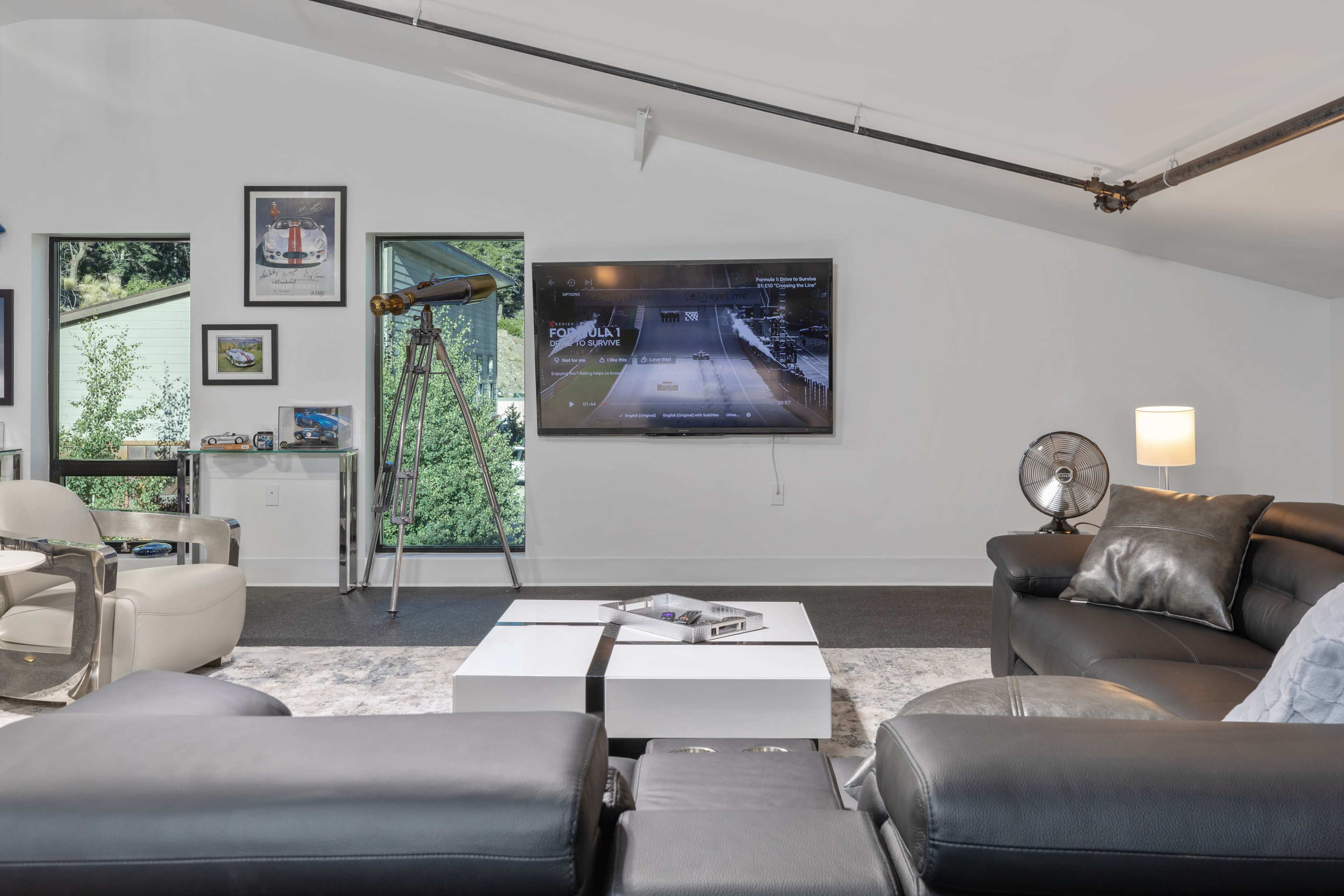 The image shows a modern living room with a large television mounted on the wall, a telescope near a window, and a combination of black leather and light-colored furniture.