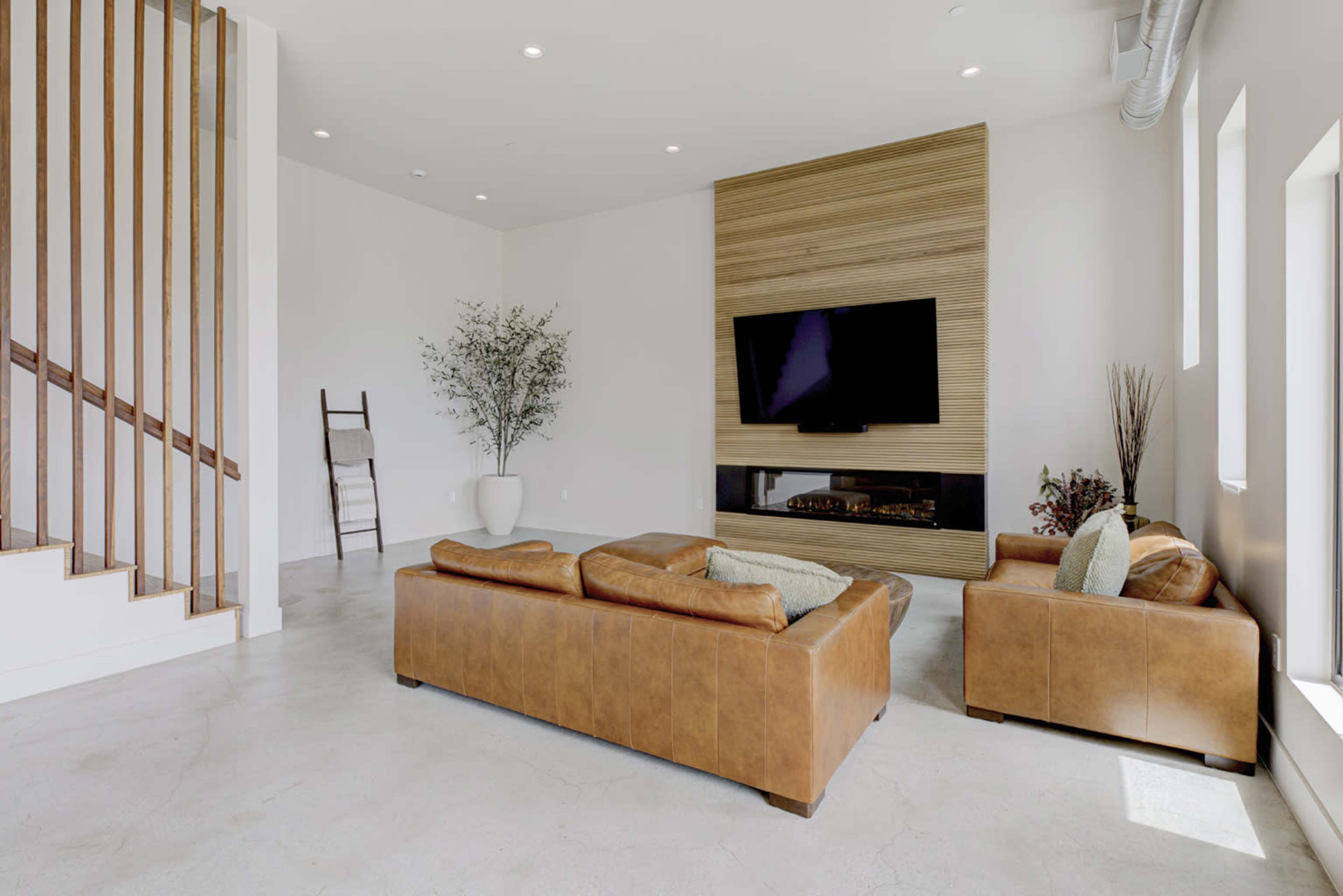 A modern living room features a brown leather sectional sofa, a flat-screen TV mounted on a wooden wall, and a decorative plant in a vase.