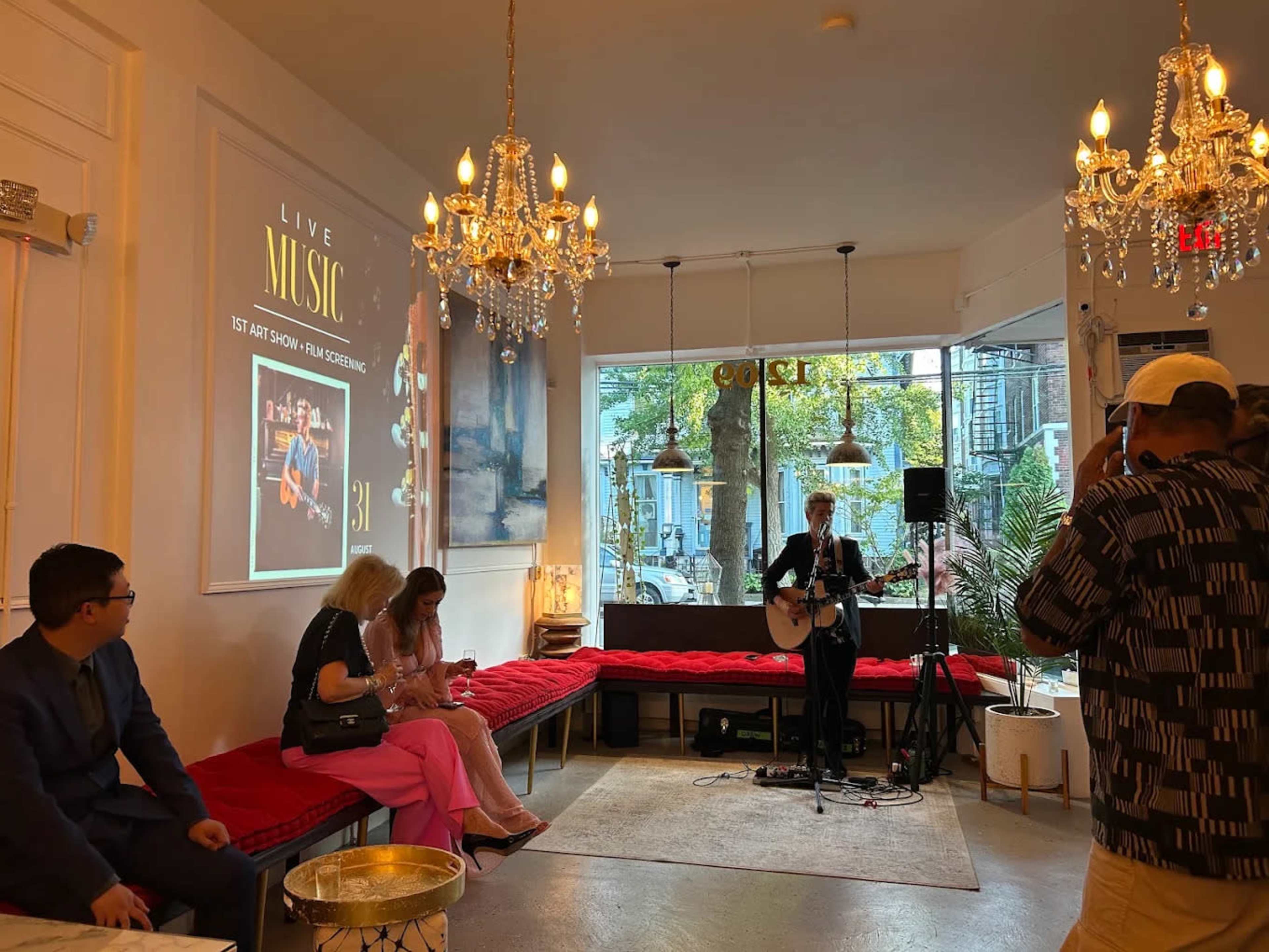 A musician performs with a guitar in a stylish lounge area featuring chandeliers and red seating, while guests watch and enjoy the live music event.