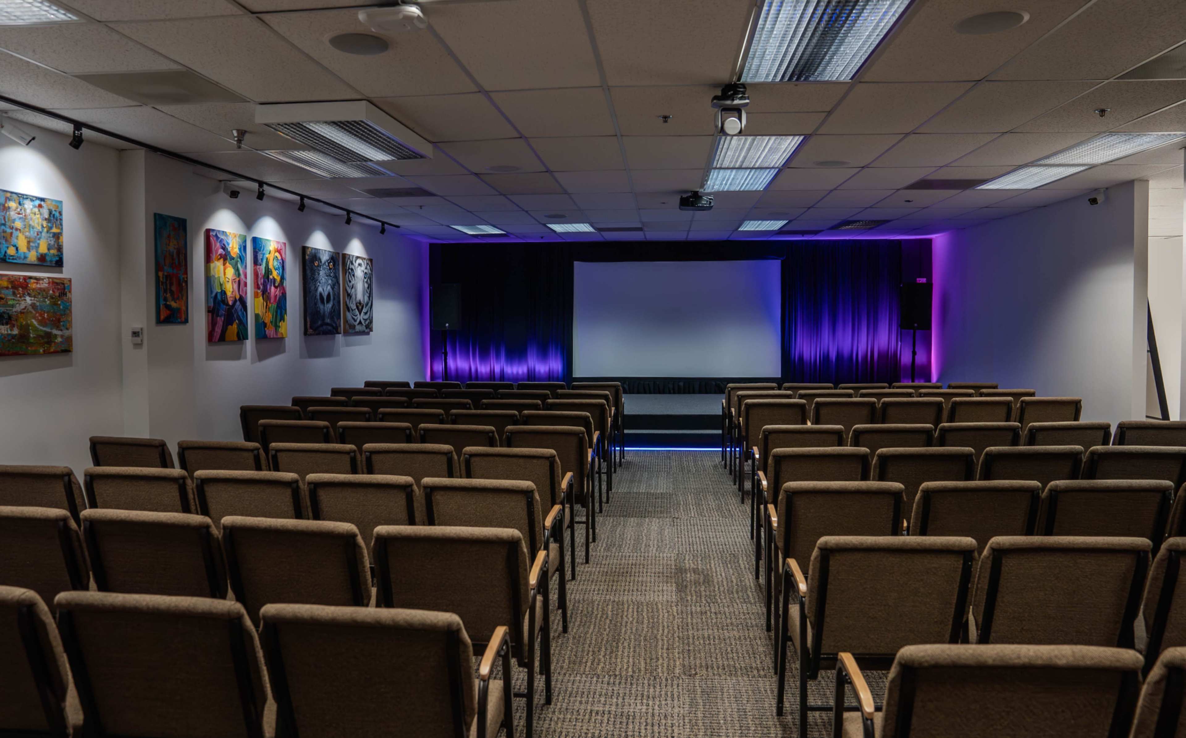The image shows a theater-style room with rows of chairs facing a central stage, which has a large screen and purple lighting in the background.