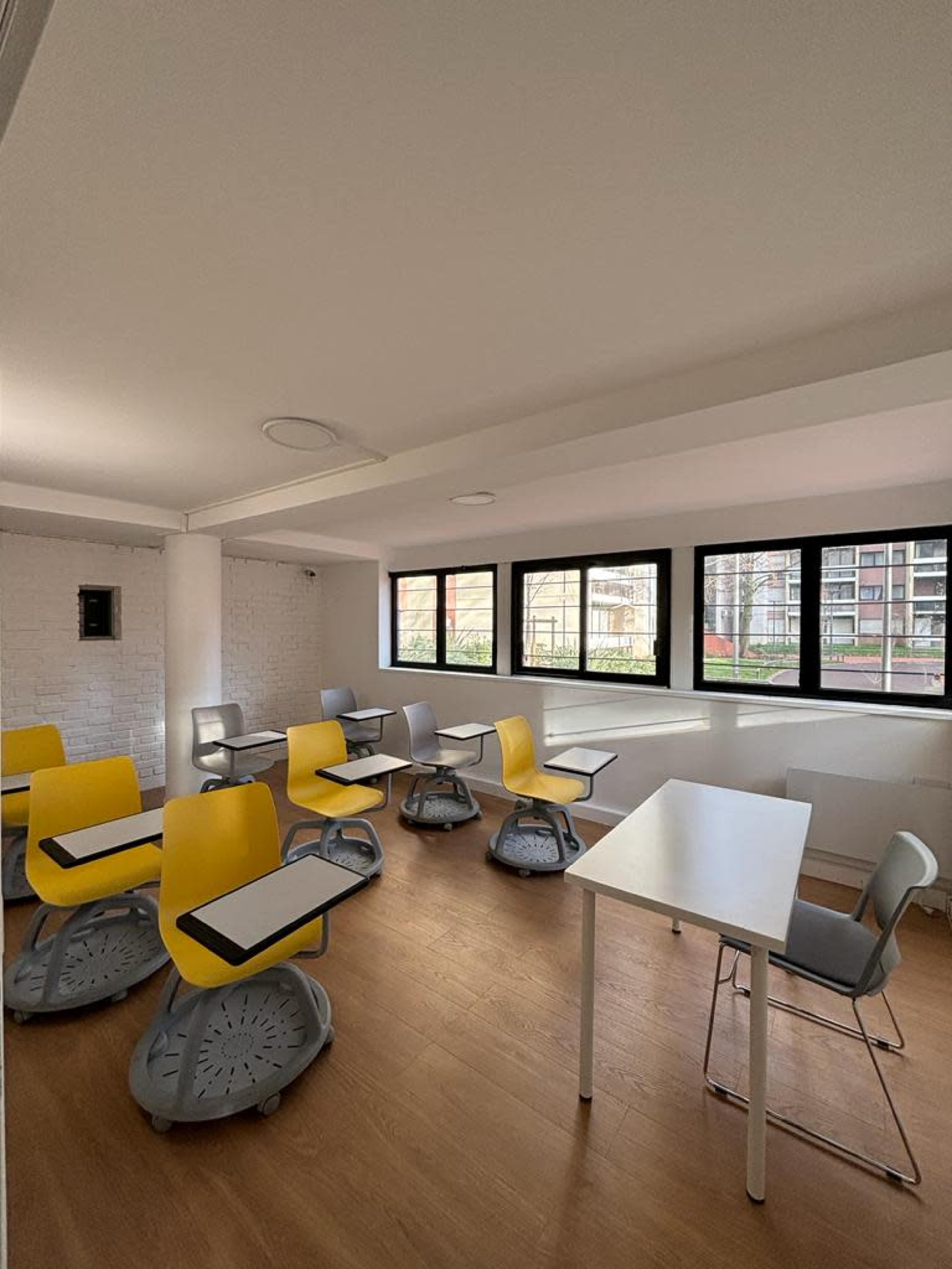 A bright classroom with several yellow and gray chairs arranged around white tables and large windows.