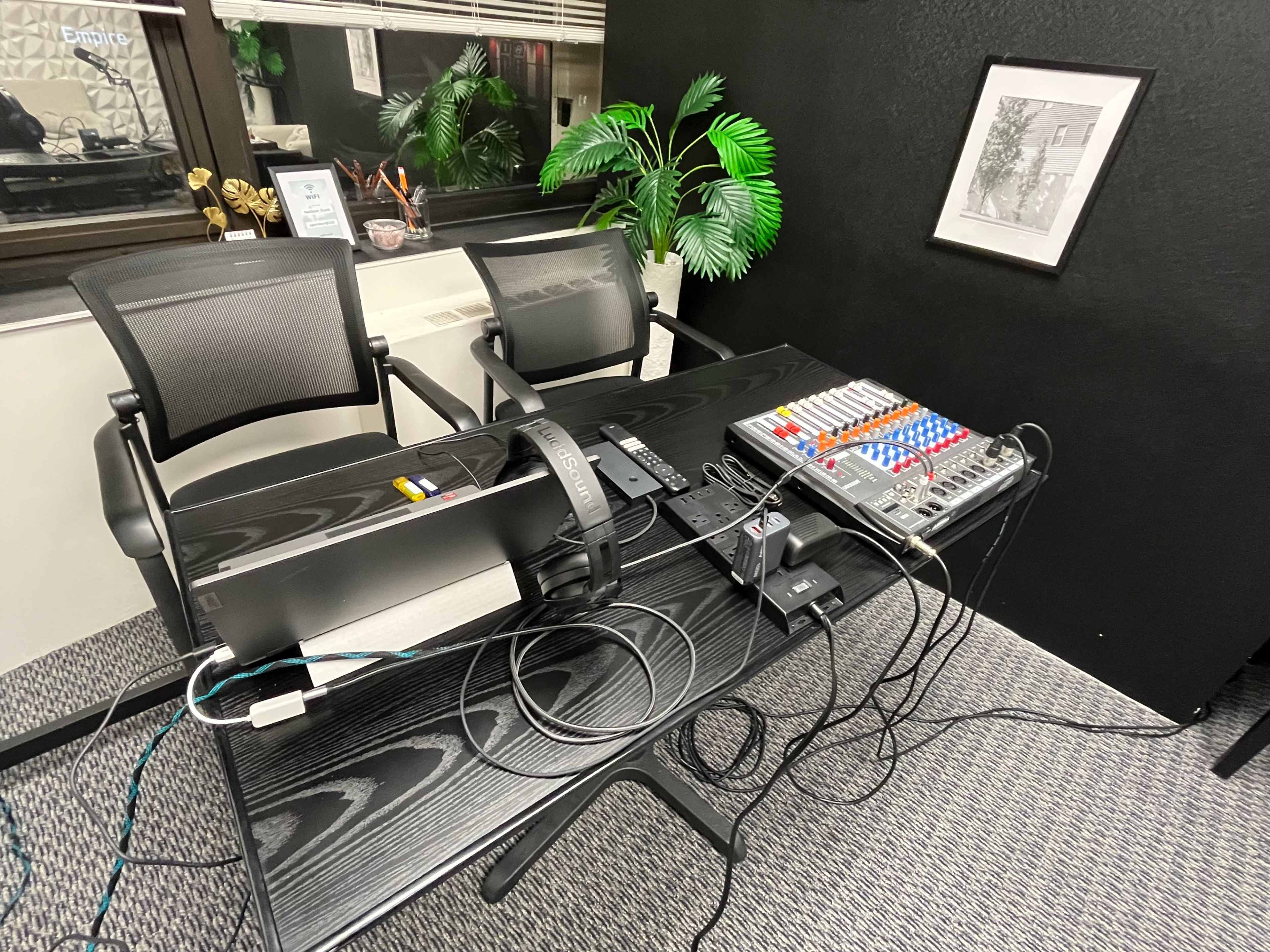 A black table in an office setting displays a sound mixer, a monitor, and various cables, with two chairs and a green plant in the background.