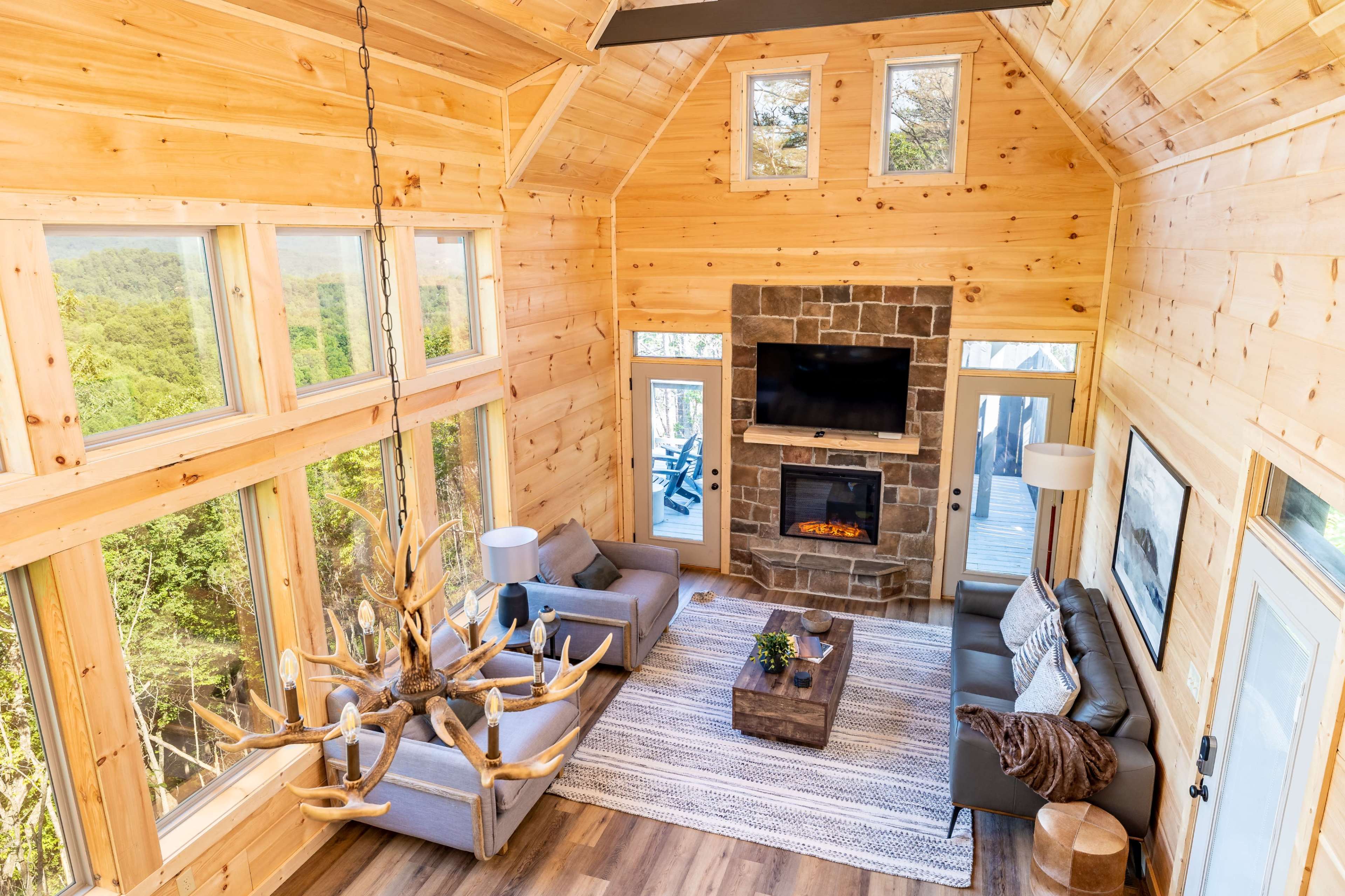 The interior of a modern cabin features tall wooden walls, a stone fireplace with a television above it, large windows that offer a view of the outdoors, and a cozy seating area with a coffee table.