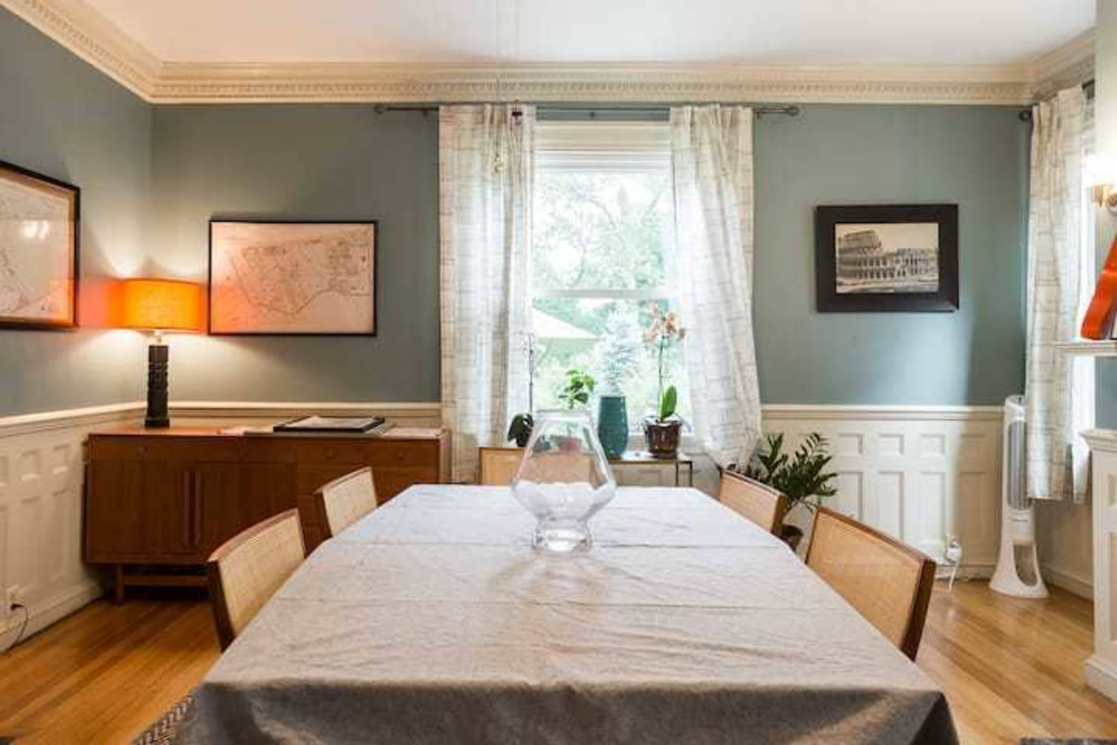 A dining room features a table at the center, with a vase on it, surrounded by chairs, and walls adorned with framed pictures.