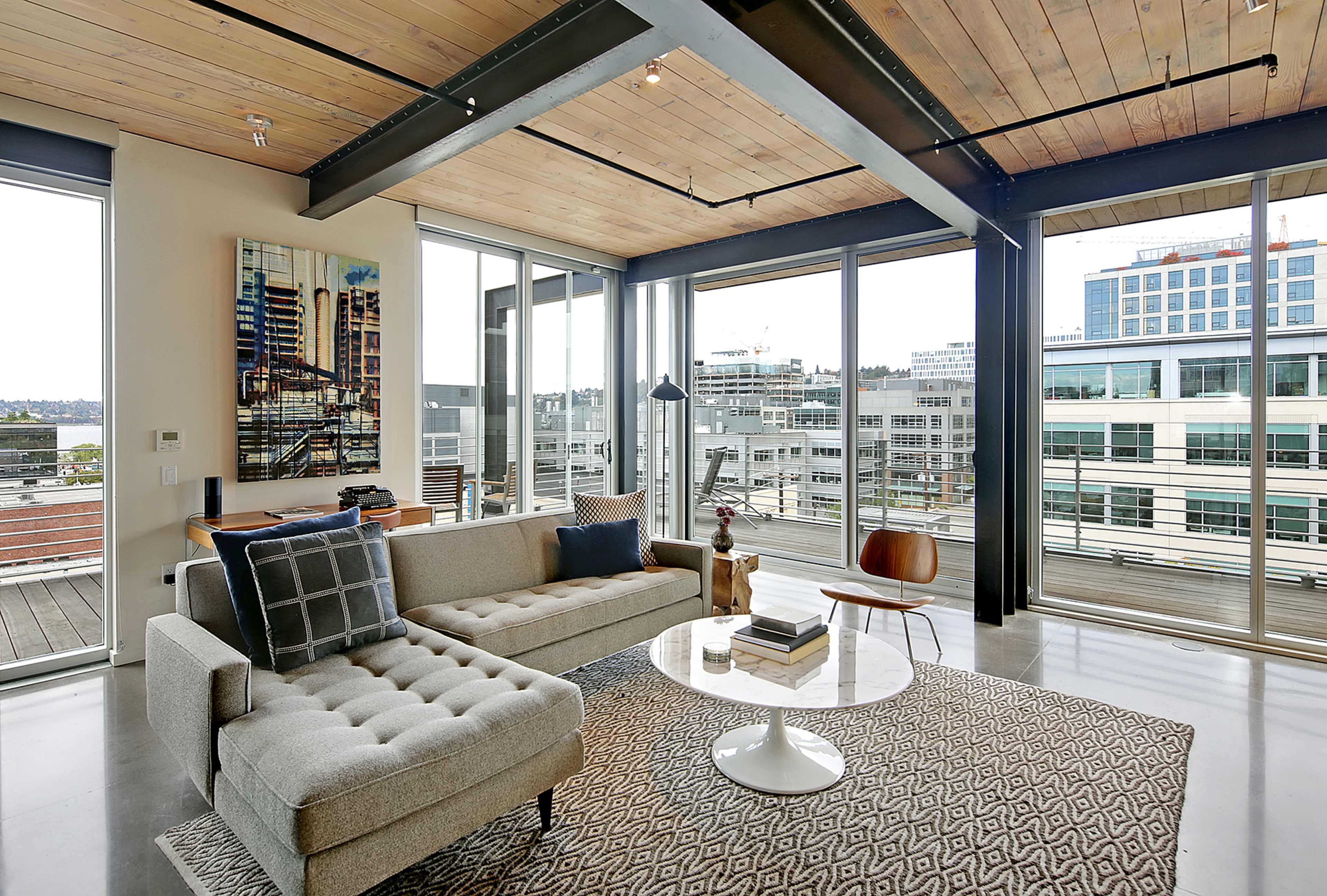 A modern living room with large windows, a sectional sofa, a circular coffee table, and a patterned rug, overlooking an urban landscape.