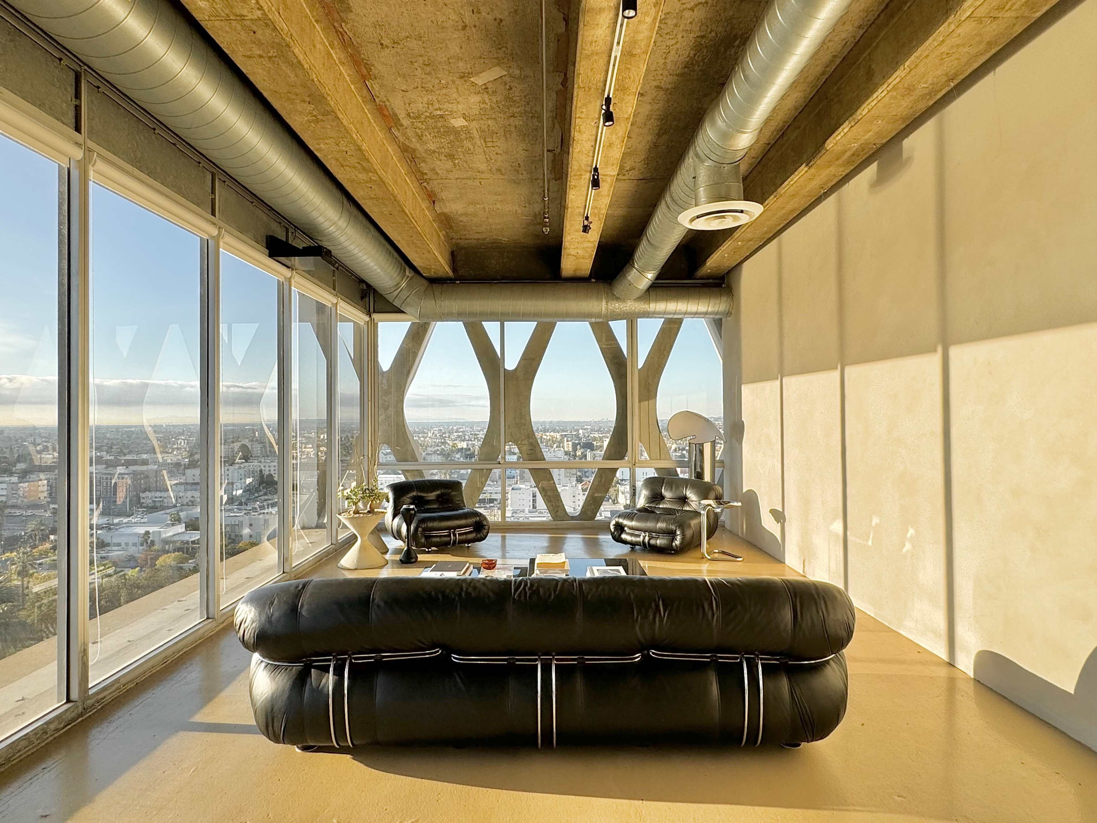 A modern living space with large windows offering a view of the city, featuring a black leather sofa and concrete ceiling beams.