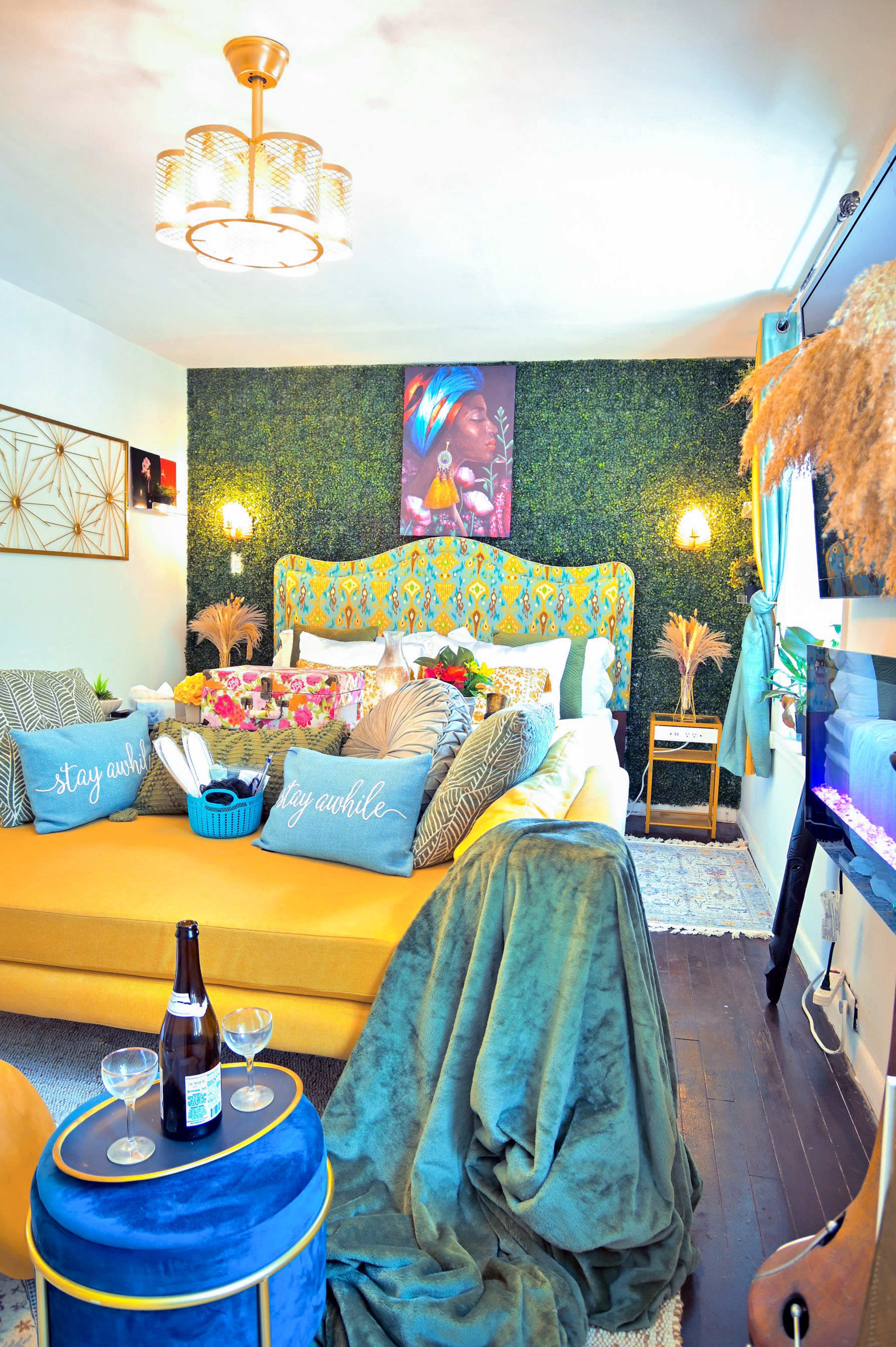 The image shows a cozy bedroom featuring a yellow bed with a colorful headboard, decorative pillows, and a green wall adorned with plants.