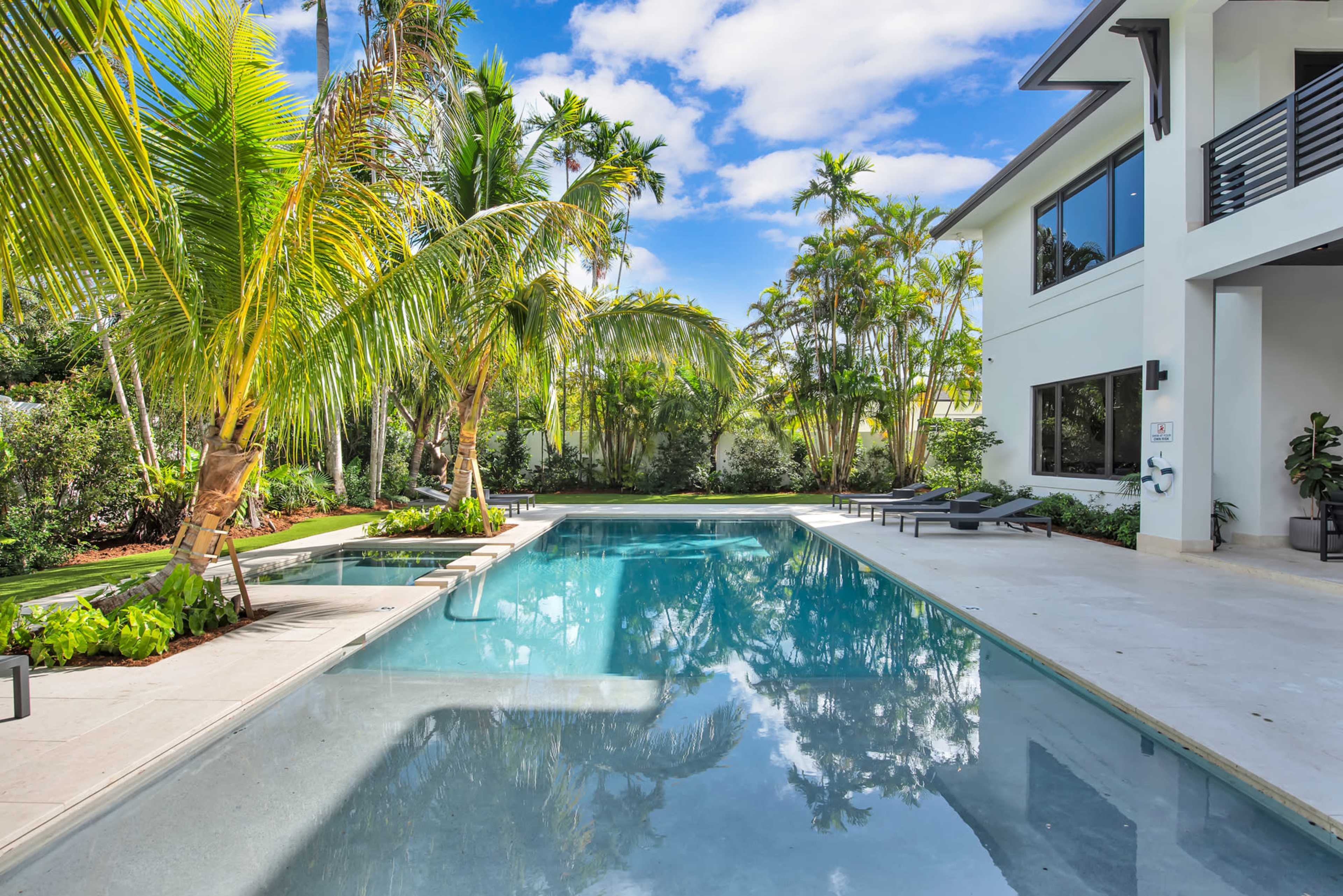 The image shows a modern home beside a clear swimming pool surrounded by tropical palm trees and lush greenery.