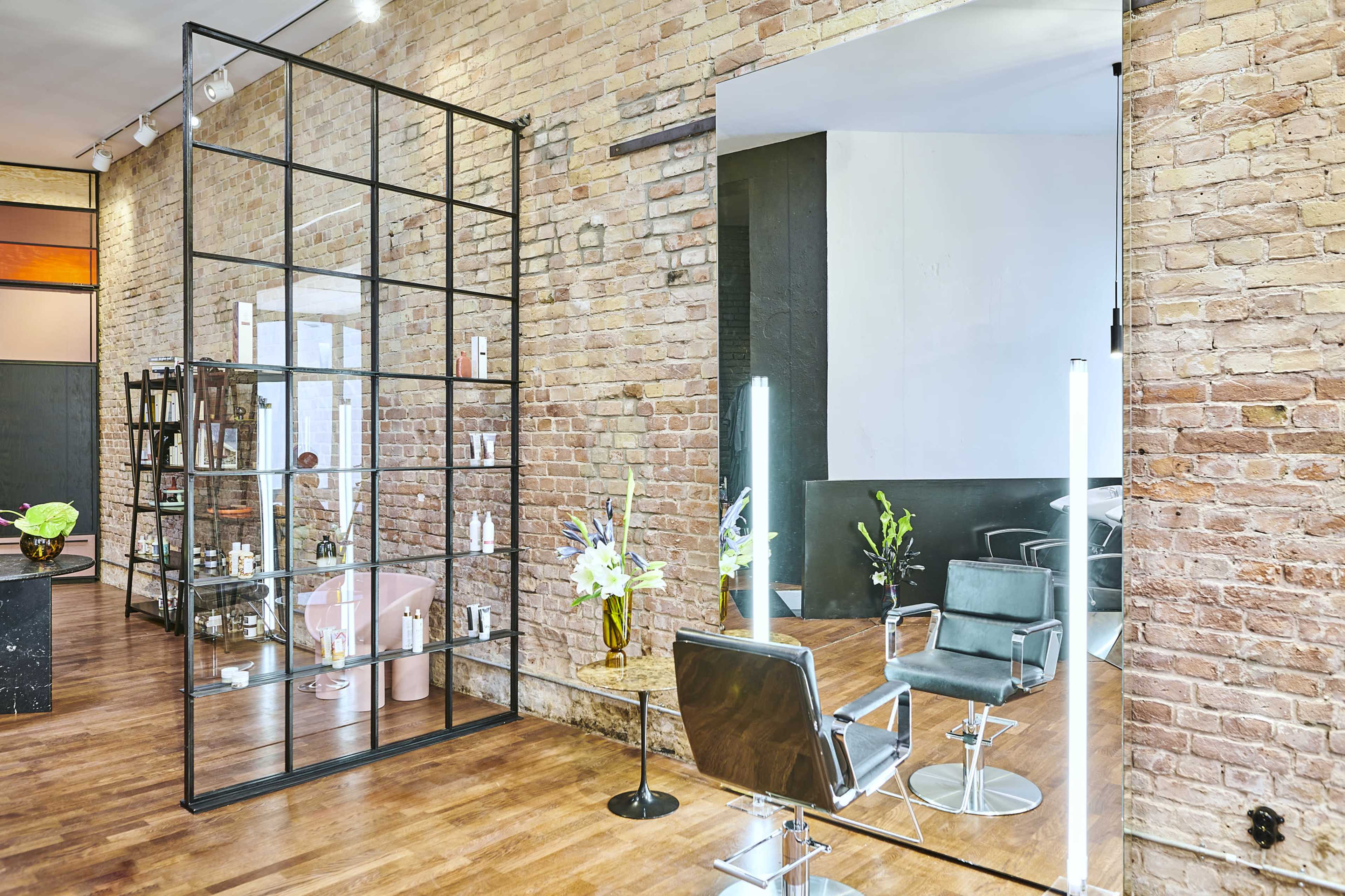 The interior of a salon features a modern design with exposed brick walls, a large mirror, and styling stations arranged alongside shelves displaying beauty products.
