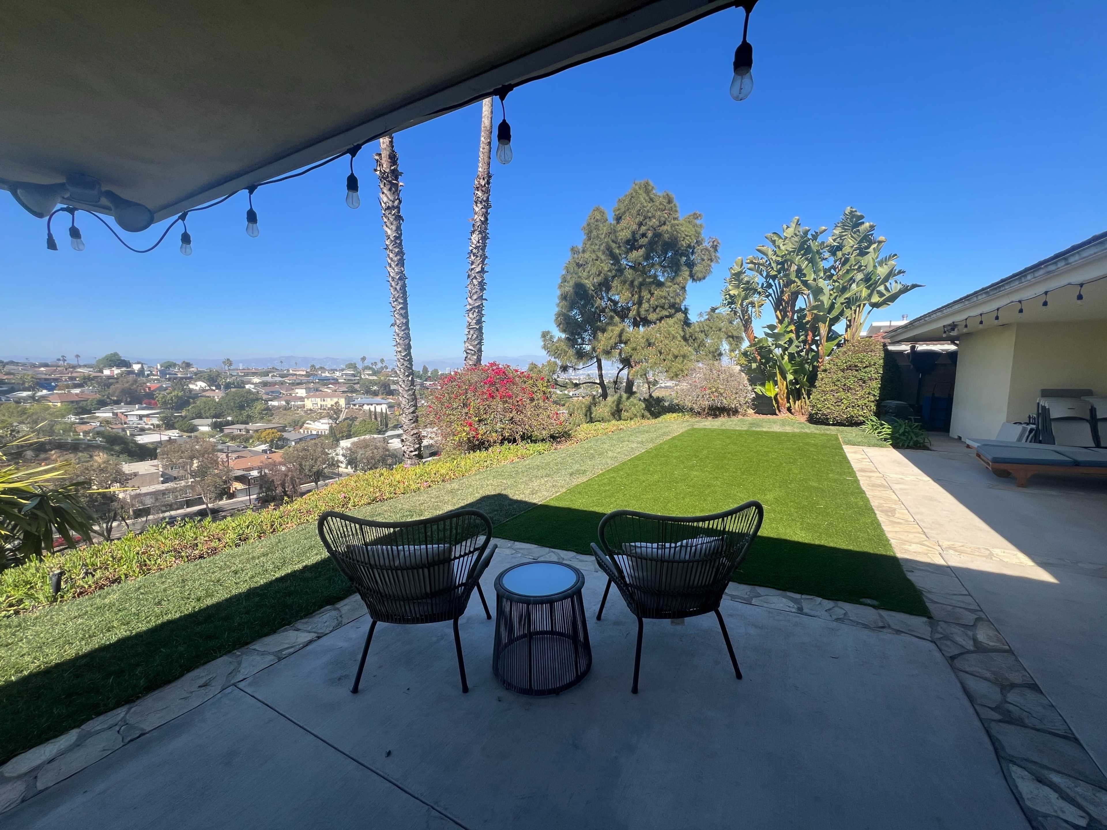 A patio with two black chairs and a small table overlooks a landscaped yard with palm trees and colorful flowers against a clear blue sky.