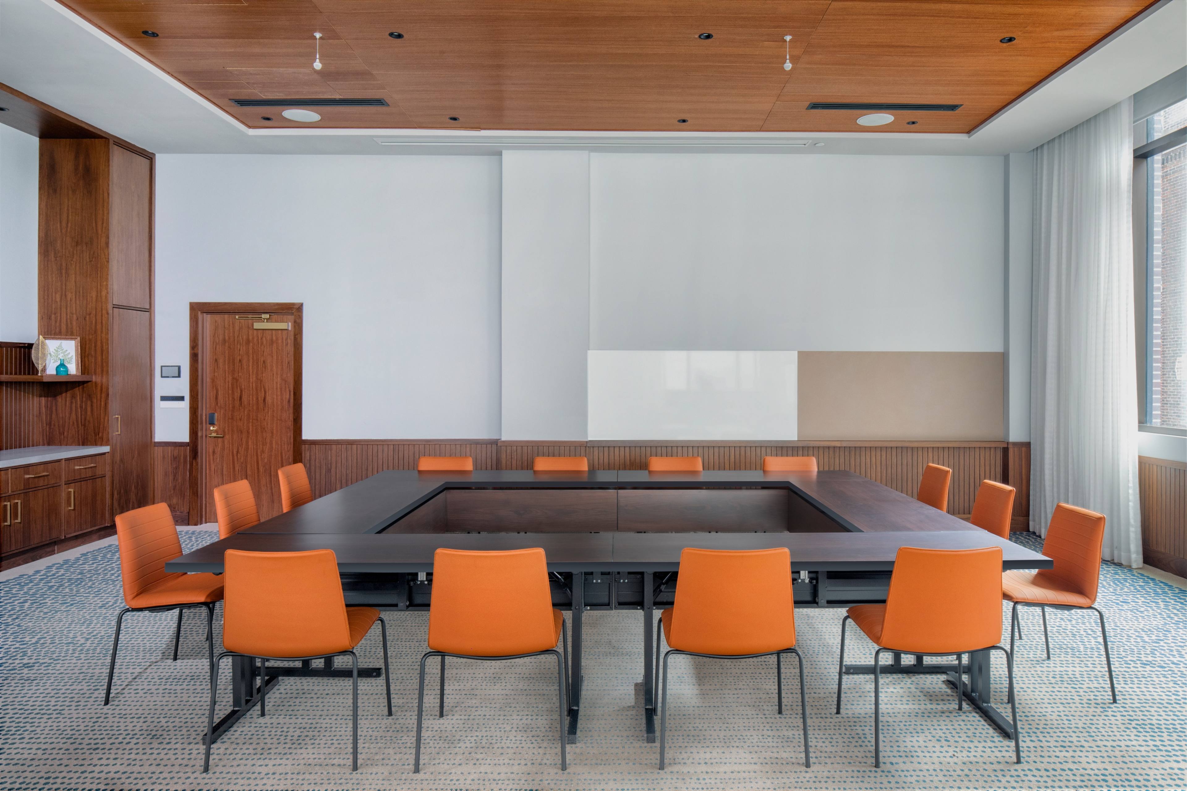 A large conference room features a dark wooden table surrounded by orange chairs on a textured carpet.