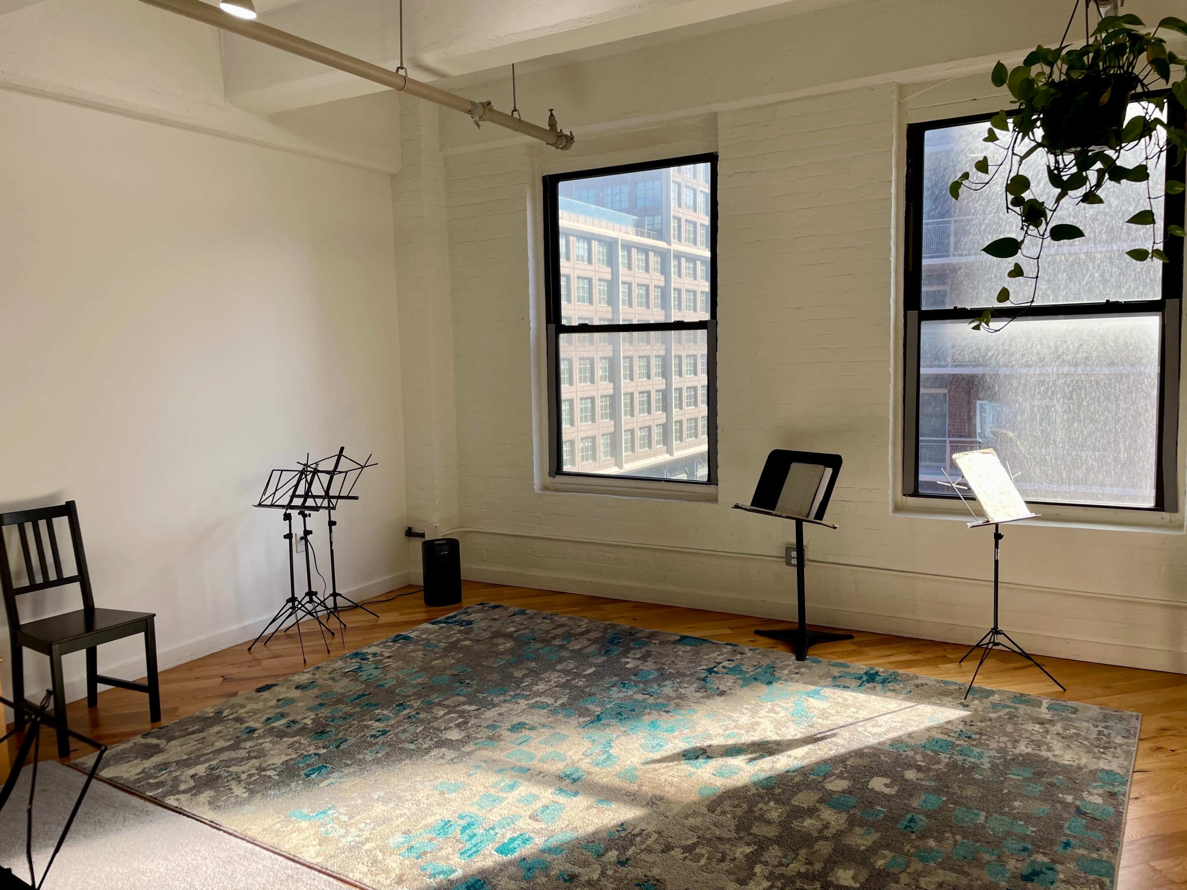 The image depicts a brightly lit room with two large windows, a patterned area rug, a chair, and three music stands positioned around the space.