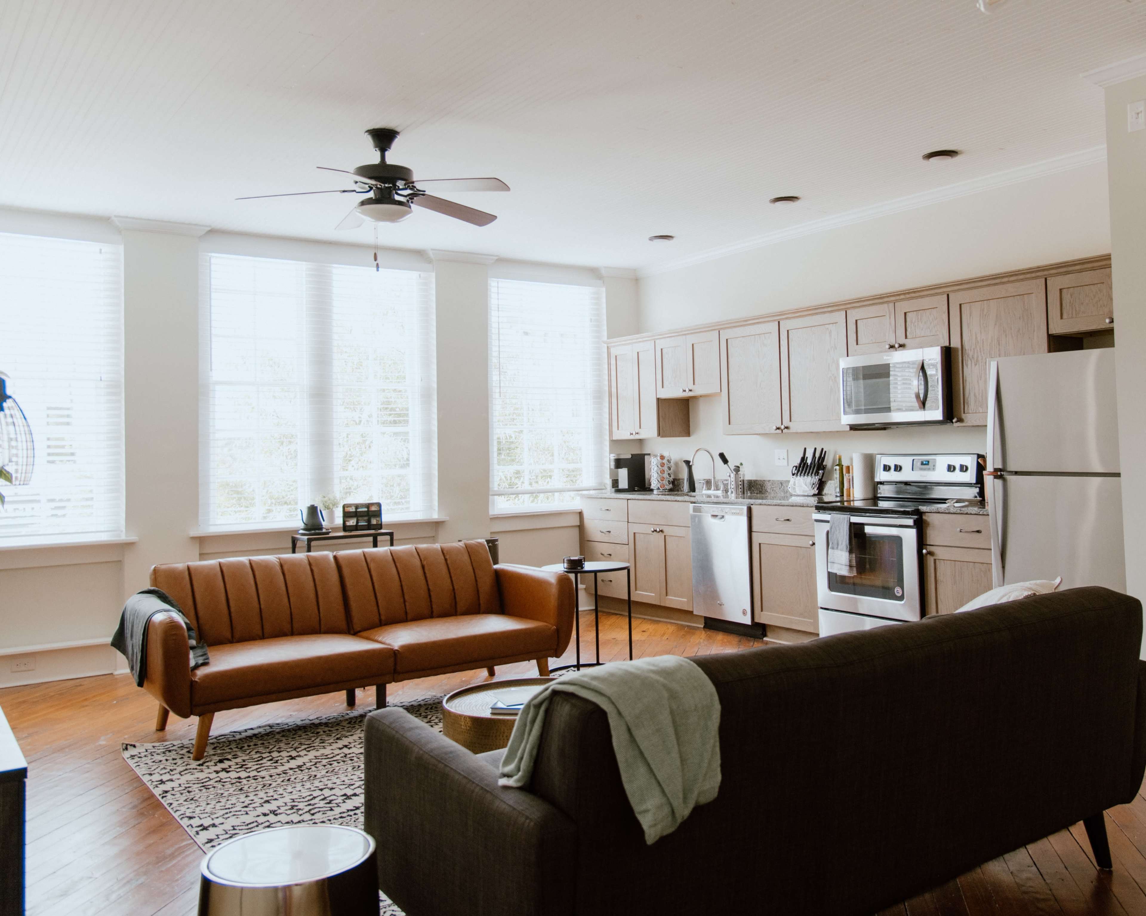 A modern living space featuring a leather sofa, a dark couch, a dining area with a round table, and a kitchen equipped with stainless steel appliances and wooden cabinets.