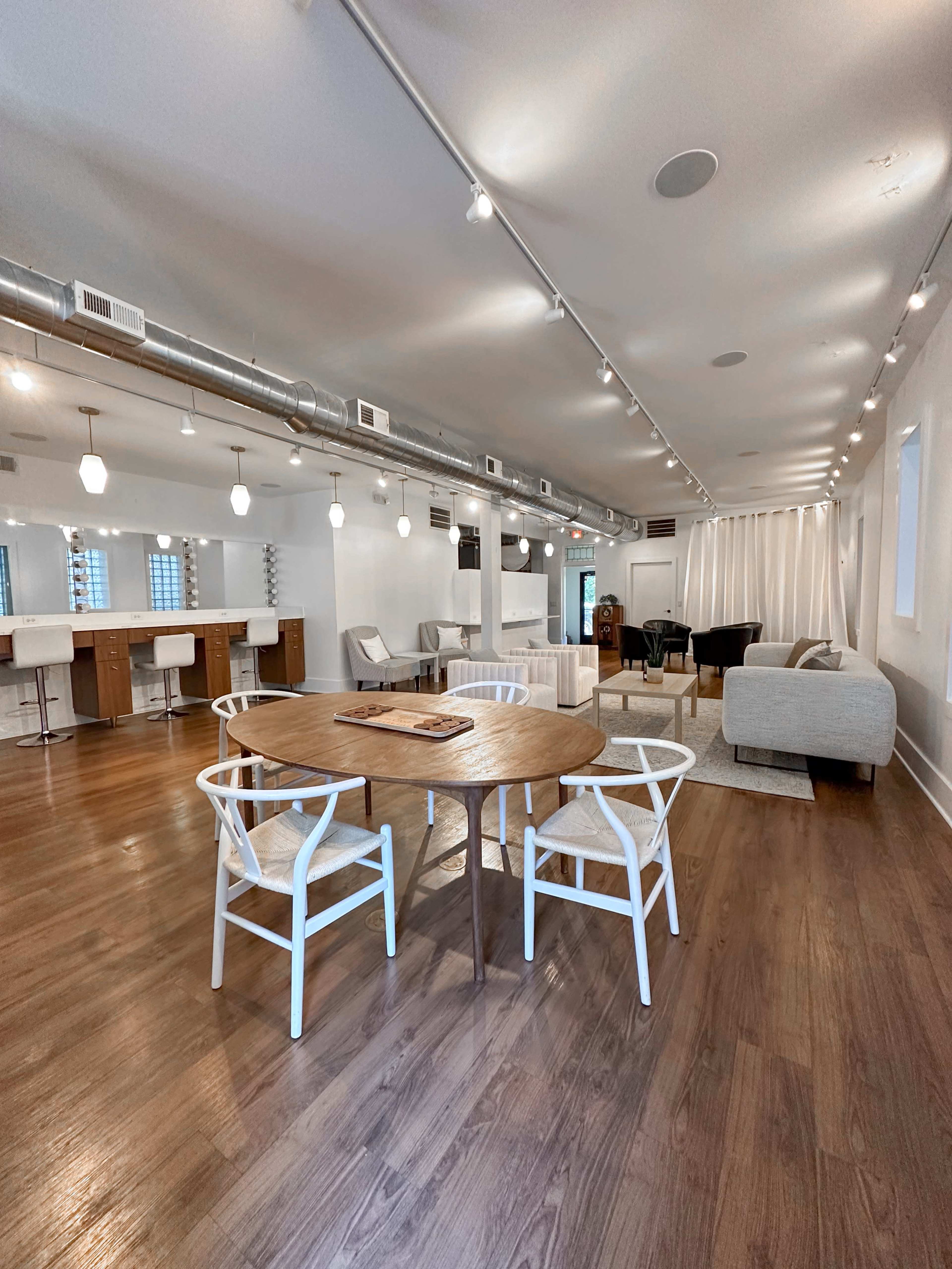 The image shows a modern, open-concept interior space featuring a round wooden dining table with white chairs, a seating area with a gray couch, and a bar counter with stools in the background.