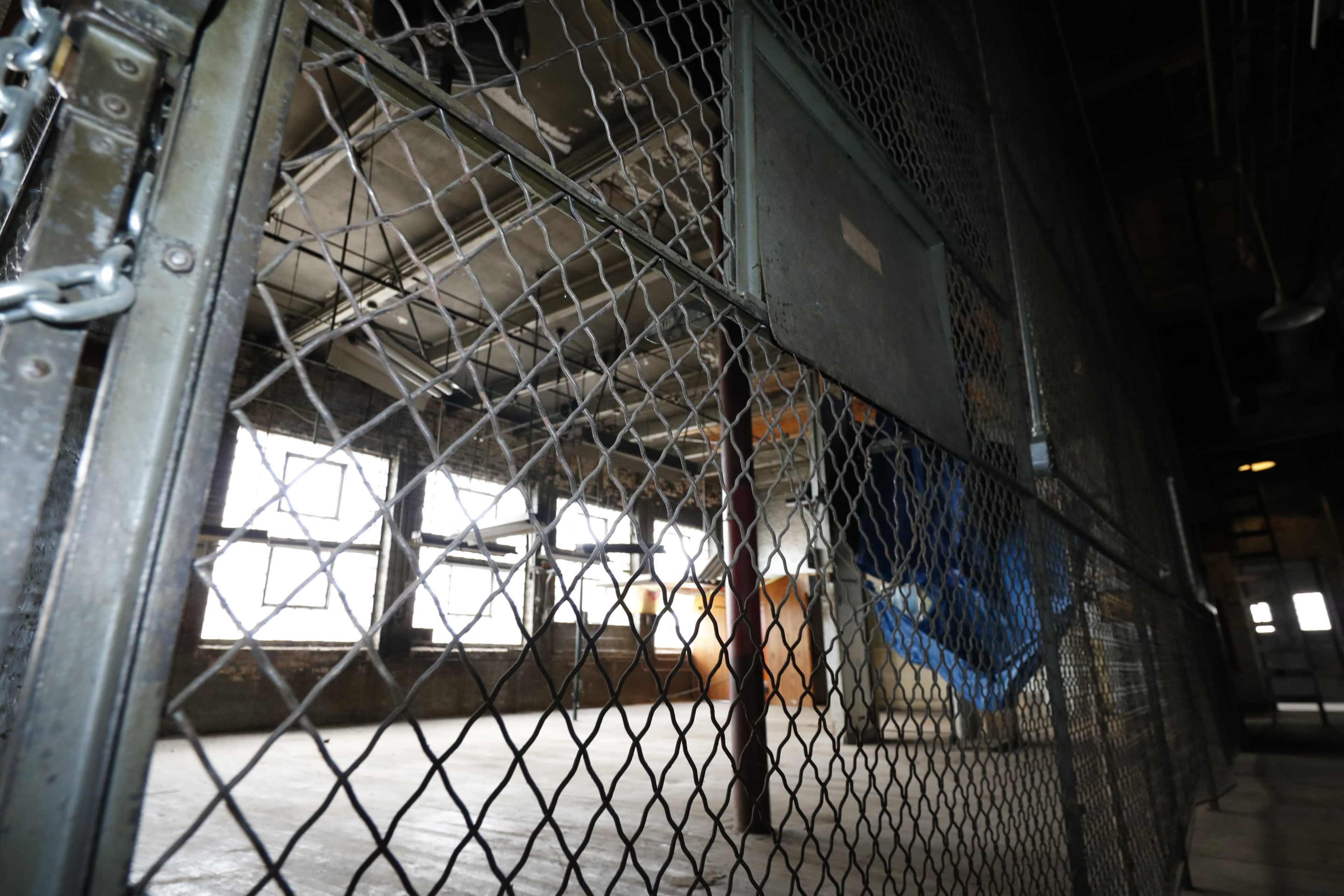 The image shows a dimly lit industrial space enclosed by a metal gate with a mesh fence and large windows in the background.