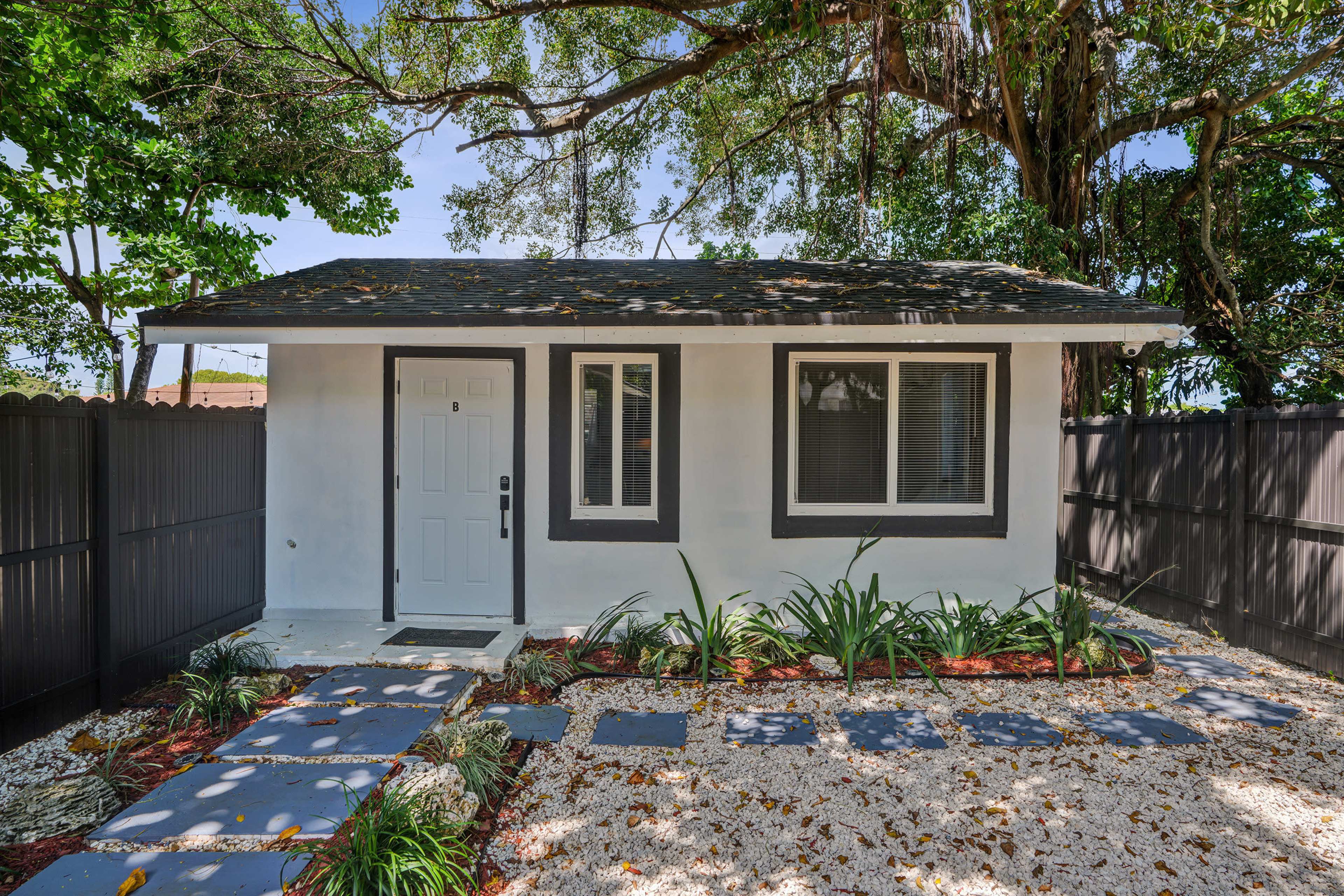 The image shows a small, modern house with a gray roof, white walls, and a stone pathway leading to the front door, surrounded by greenery and a black fence.
