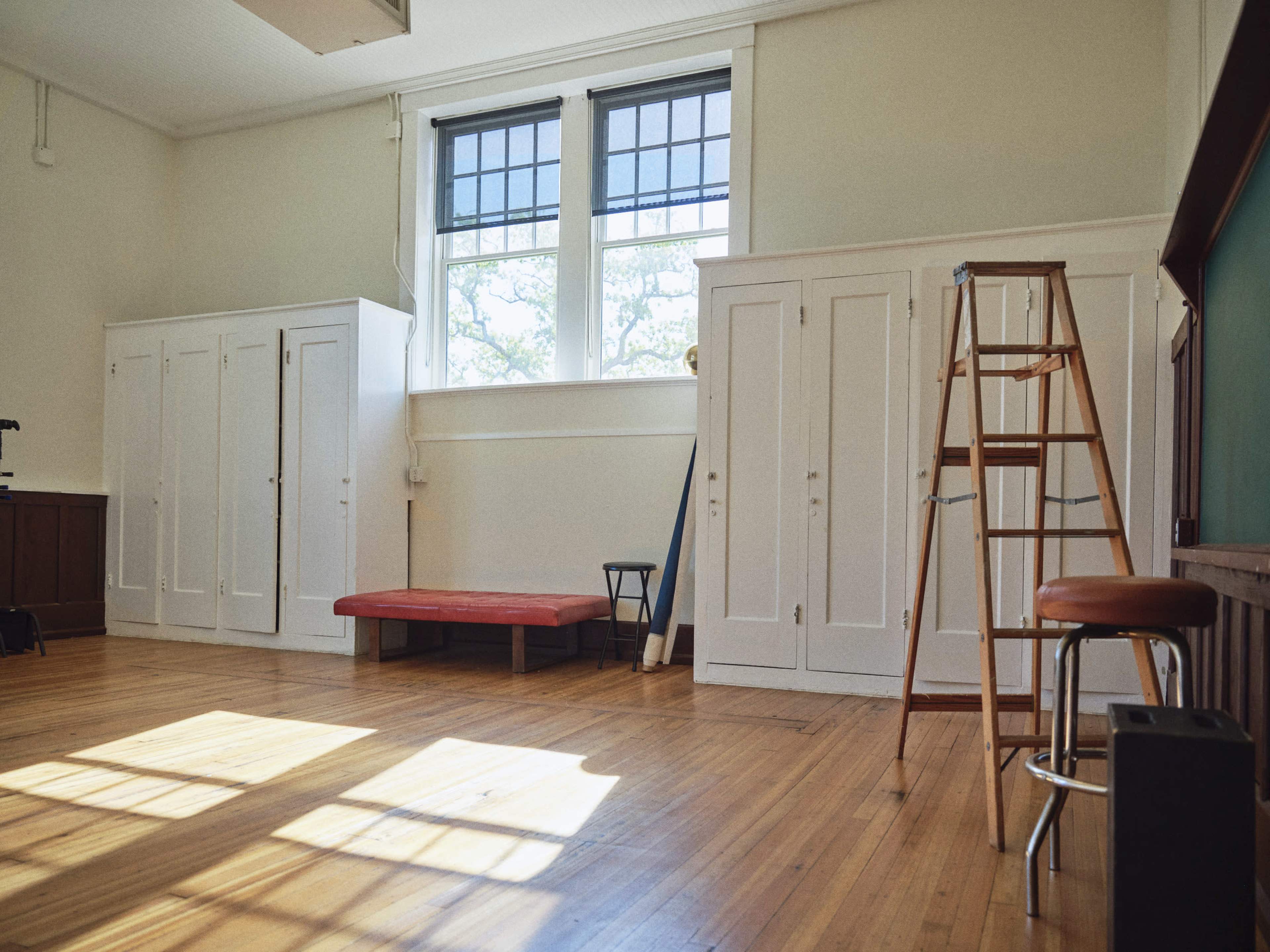 The image shows a bright room with wooden flooring, two white wardrobes, a red bench, a ladder, and a bar stool near a green chalkboard.