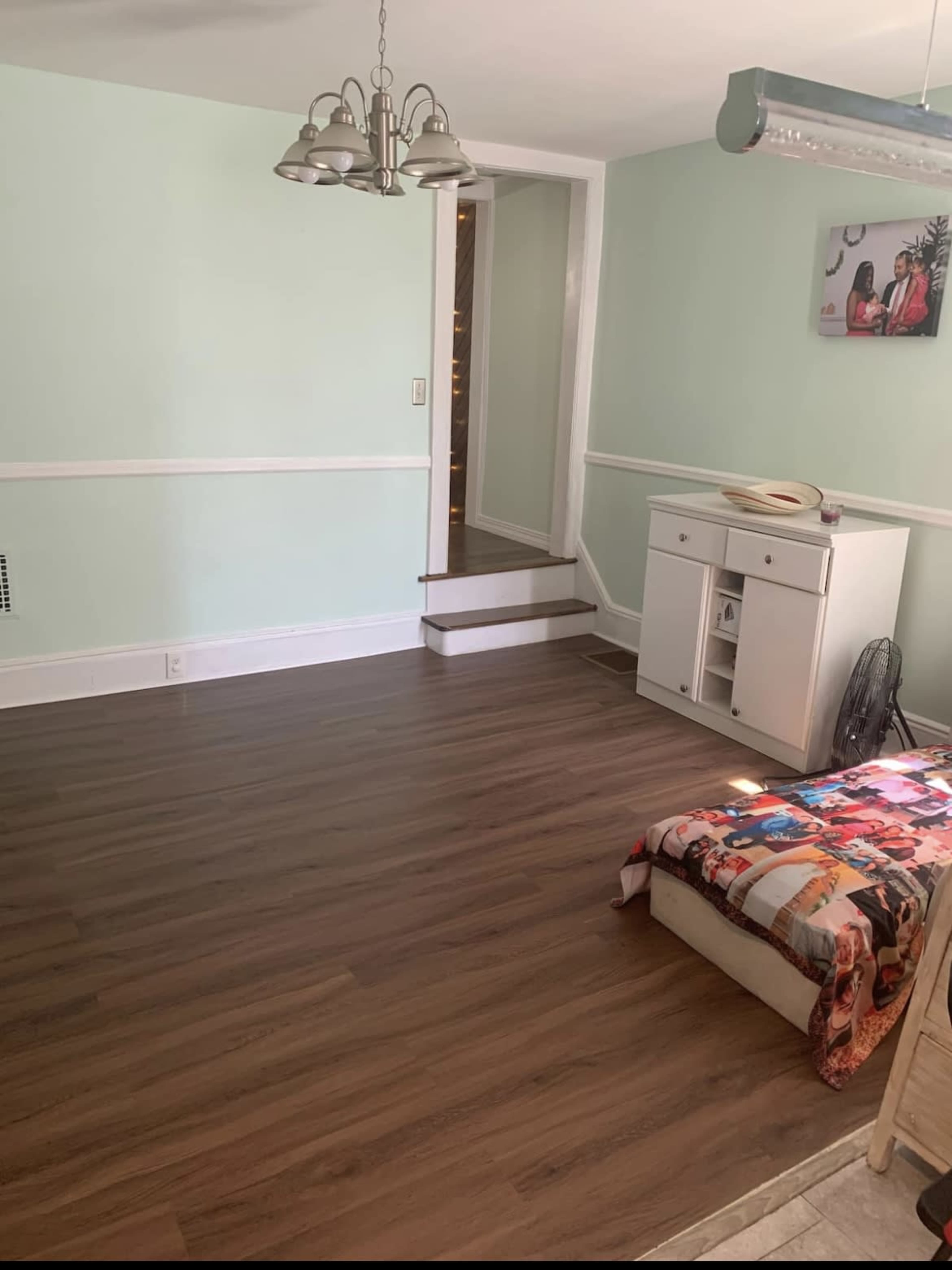 A room with light green walls, a wooden floor, a small table with a chair, and a bed against one wall.