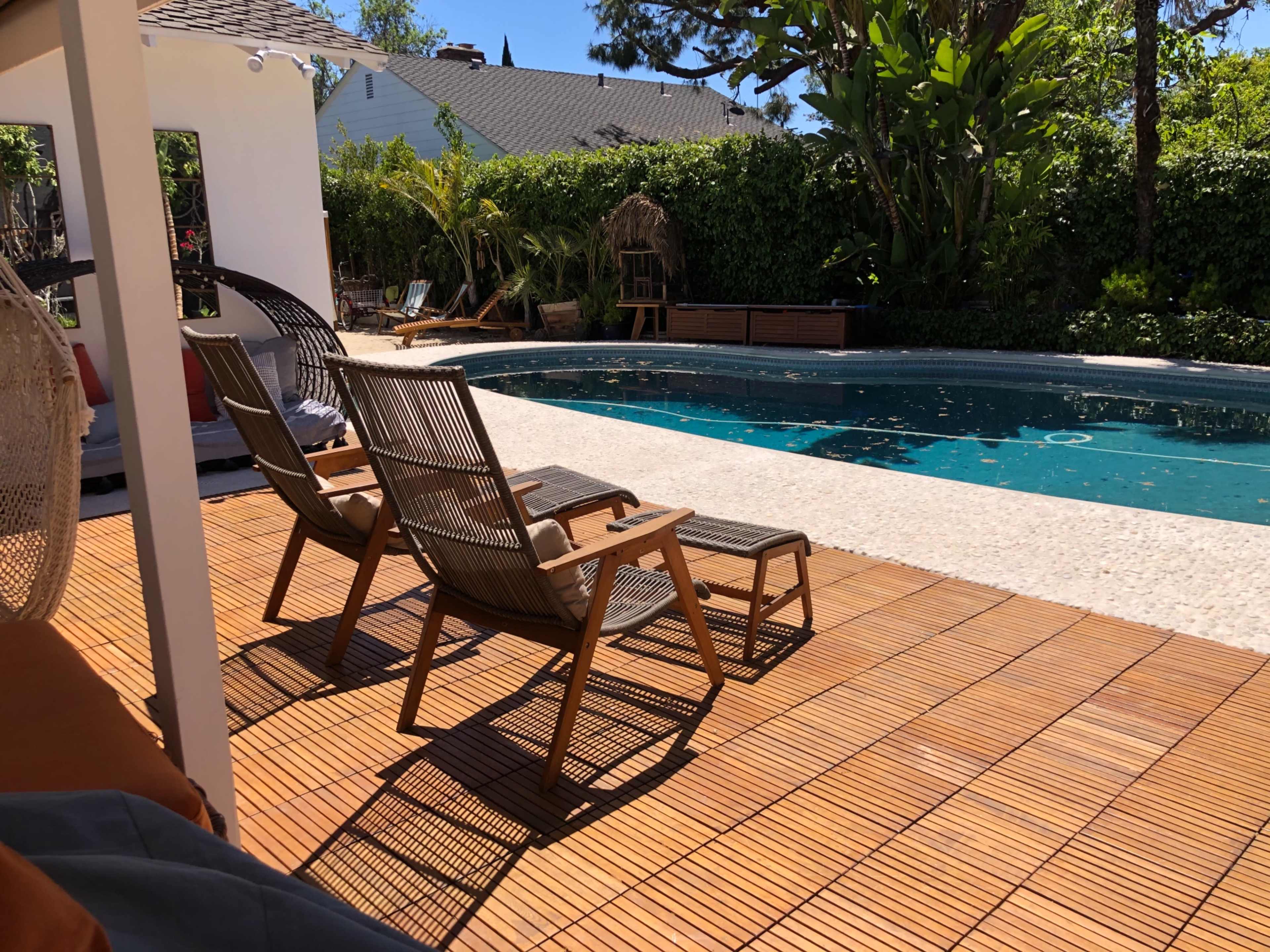 A wooden deck with two reclining chairs overlooks a swimming pool surrounded by greenery.