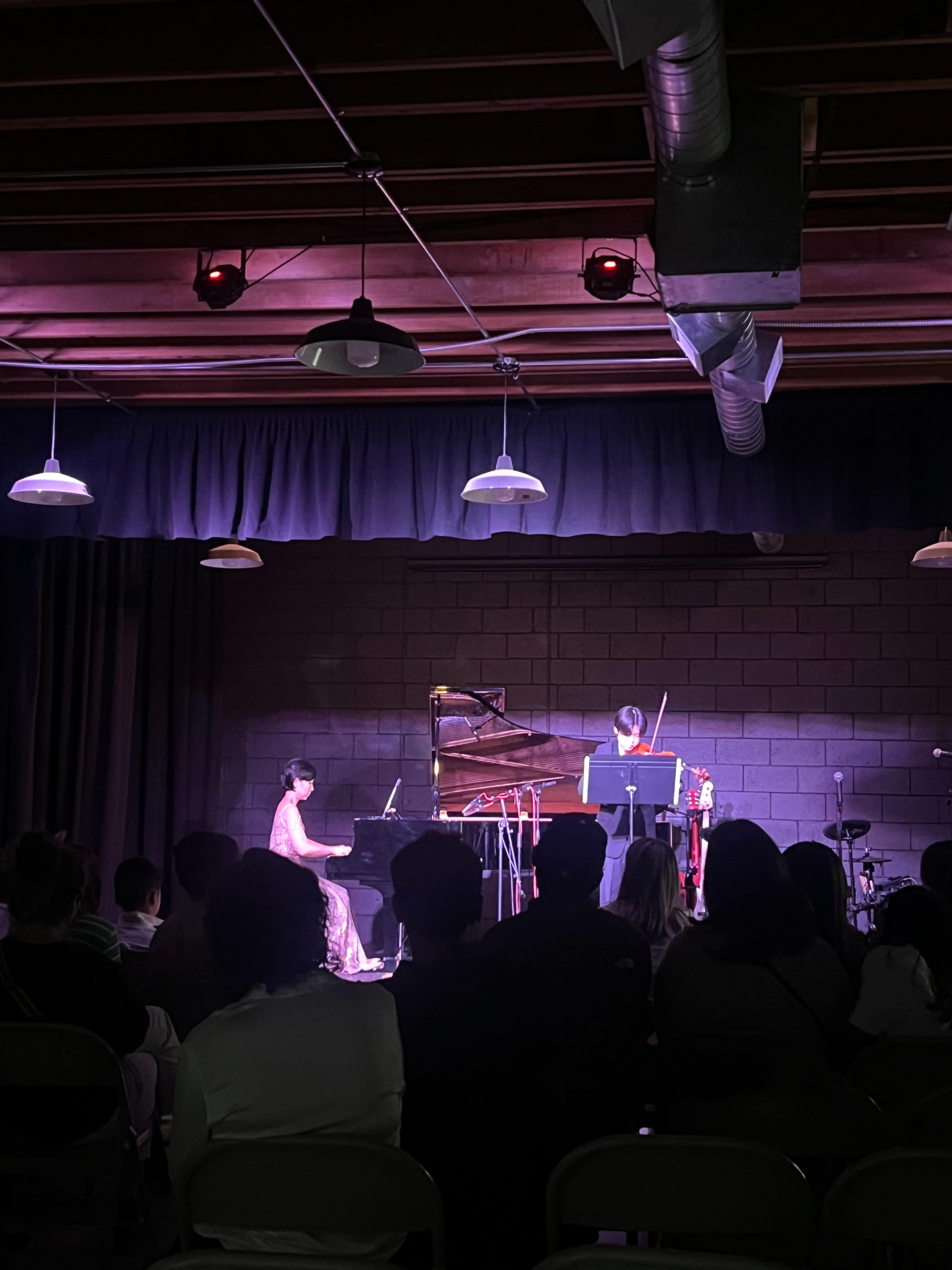 A pianist and a violinist perform on stage in a dimly lit venue with an audience seated in front.