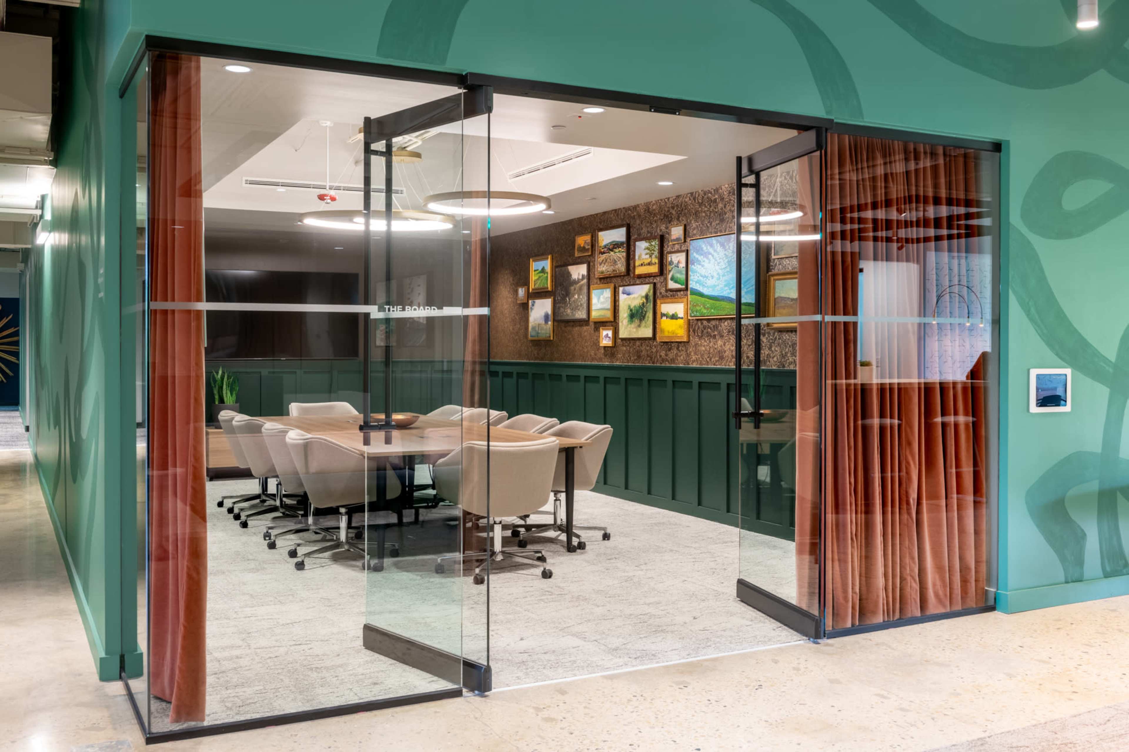 A glass-walled meeting room with a round table and chairs, surrounded by green walls and artwork.