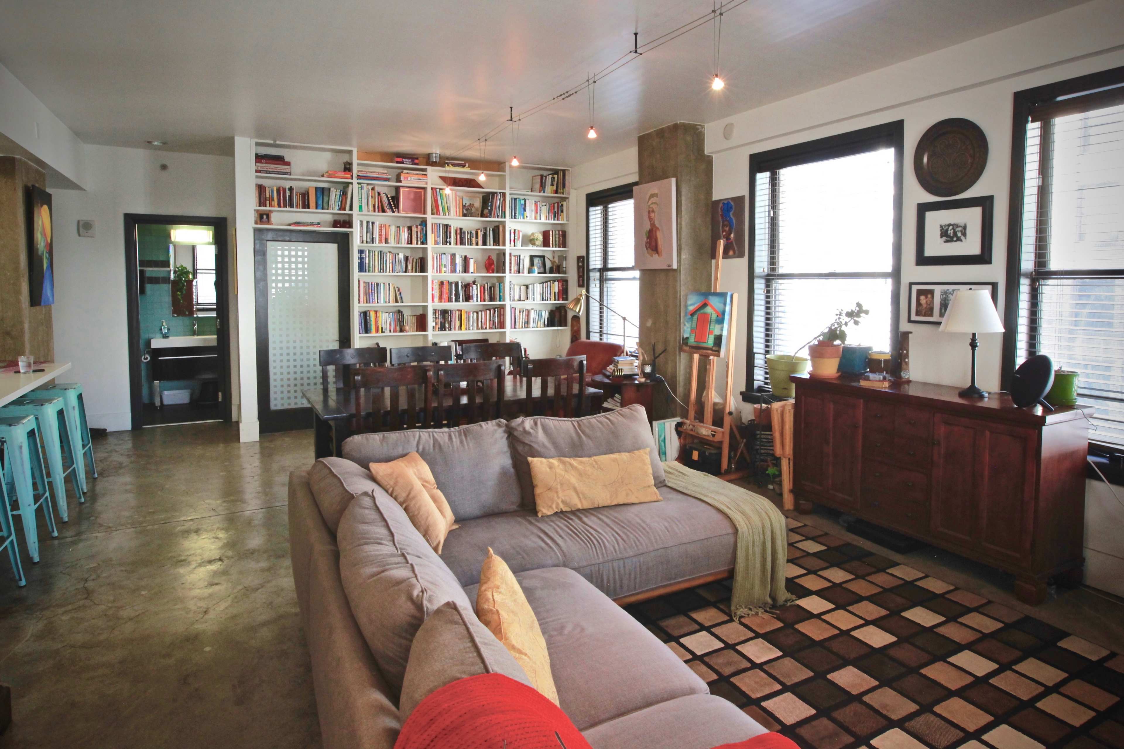 The image shows a modern living room with a gray sectional sofa, a large bookshelf filled with books, and an adjacent dining area with teal bar stools and a wooden dining table.