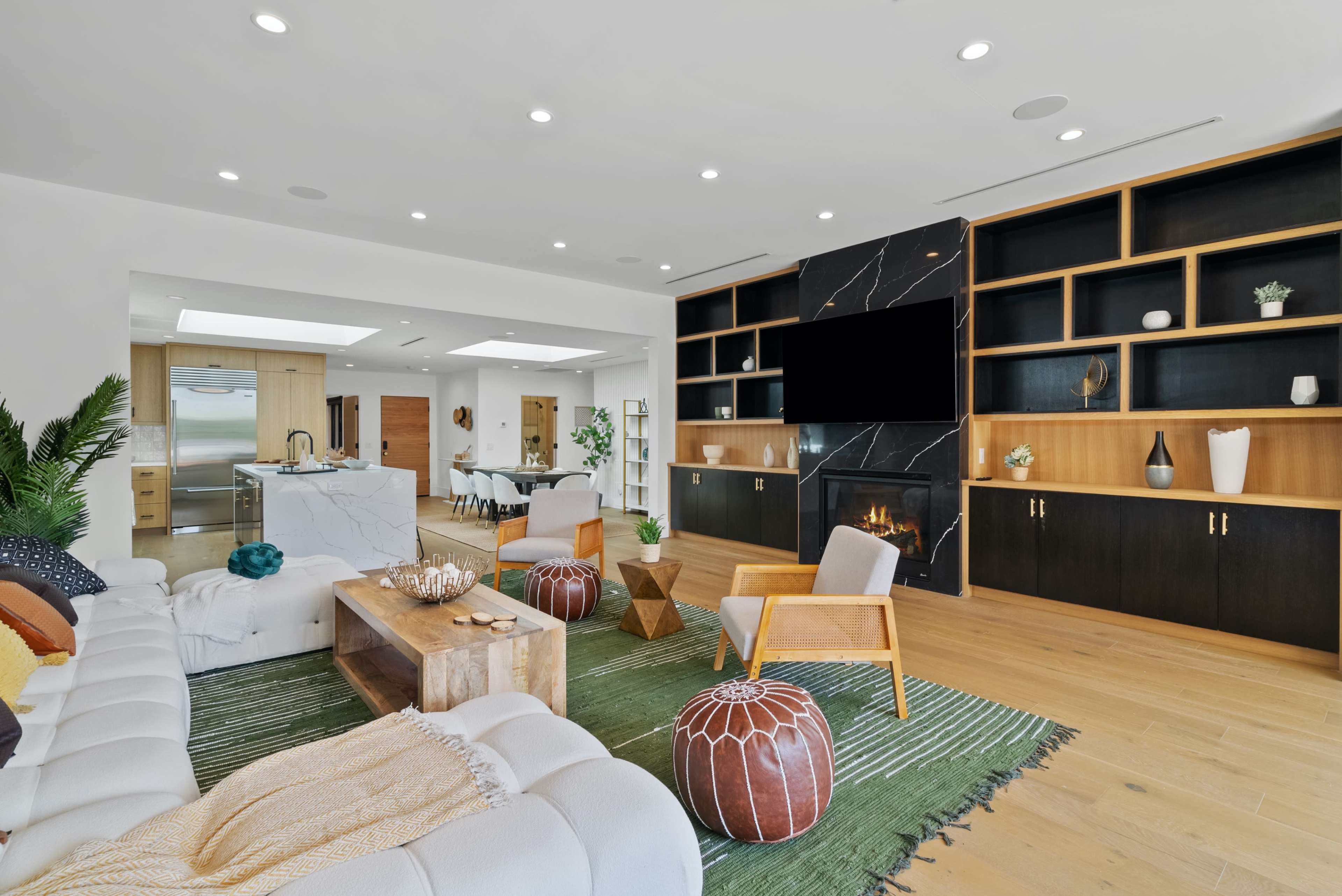 The room features a modern living area with a white sectional sofa, wooden coffee table, and a black marble media wall with a fireplace, surrounded by shelves and a green area rug.