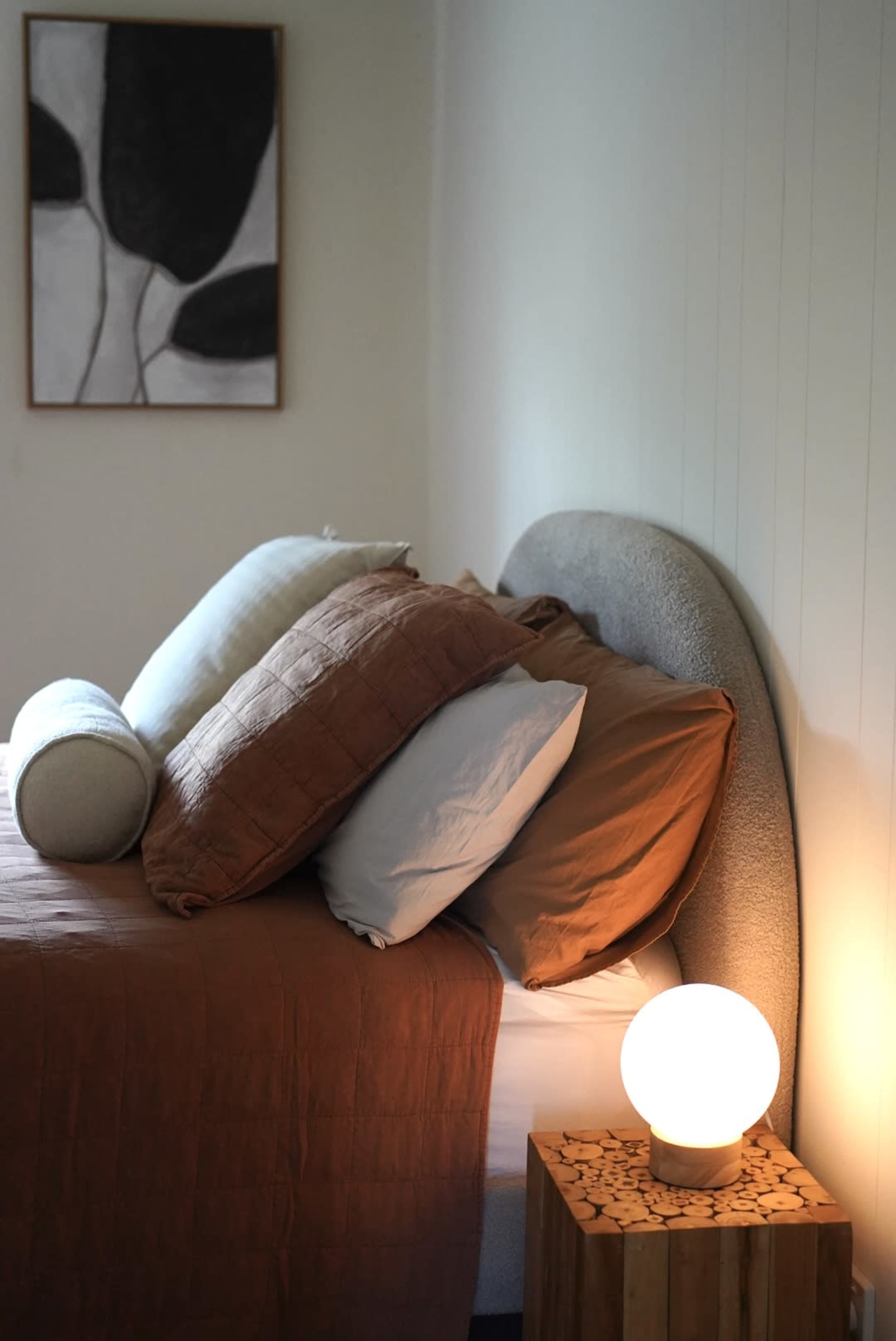 The image shows a neatly made bed with multiple pillows and a lamp beside it, along with a framed artwork on the wall.