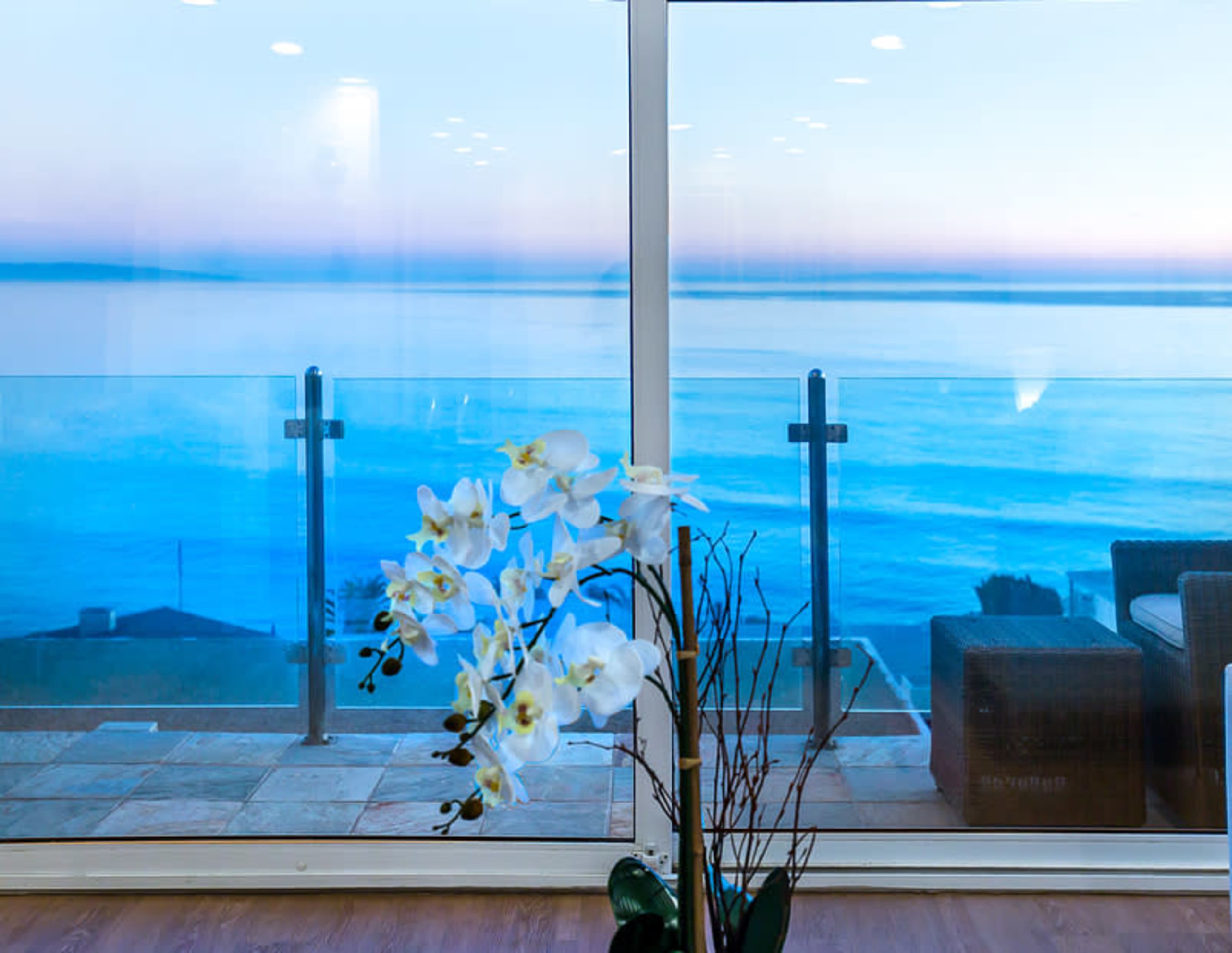 A white orchid plant sits in the foreground of a large glass window overlooking the calm ocean at dusk.