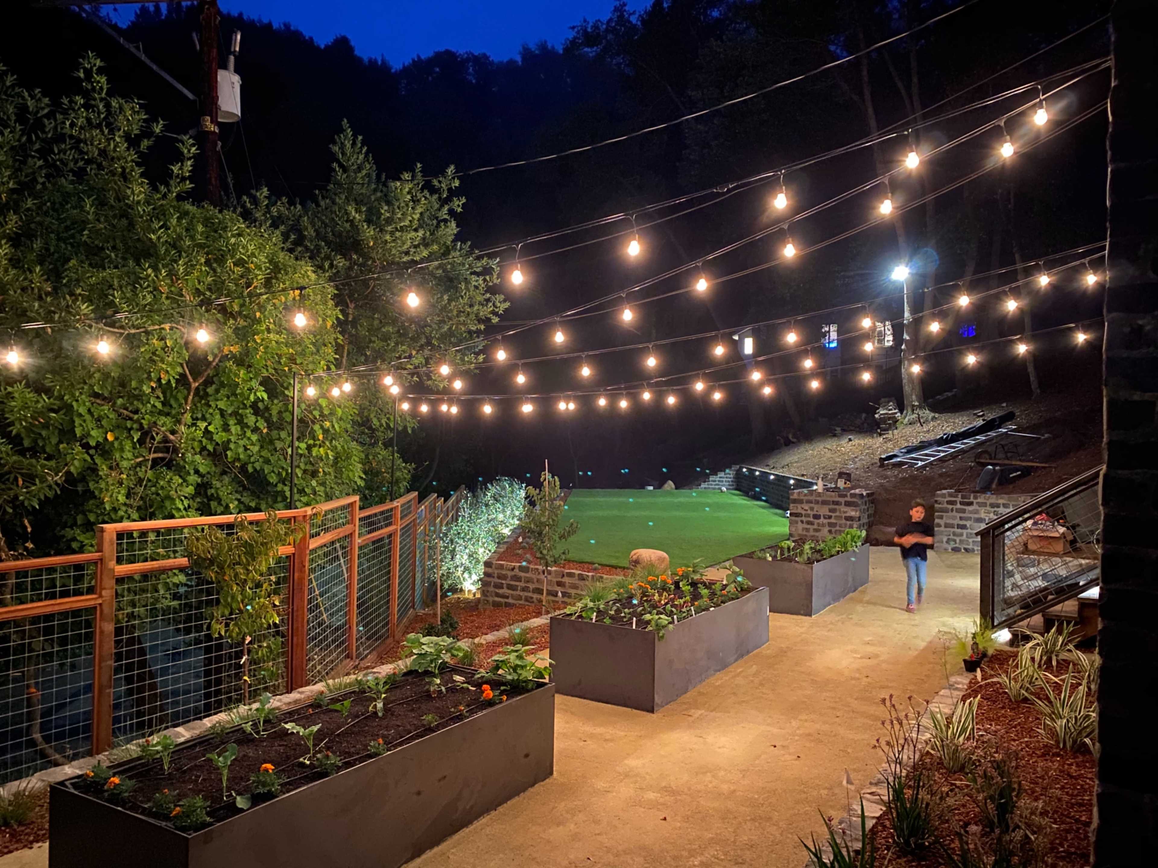 A pathway lined with string lights and raised garden beds leads toward a grassy area surrounded by trees at dusk.