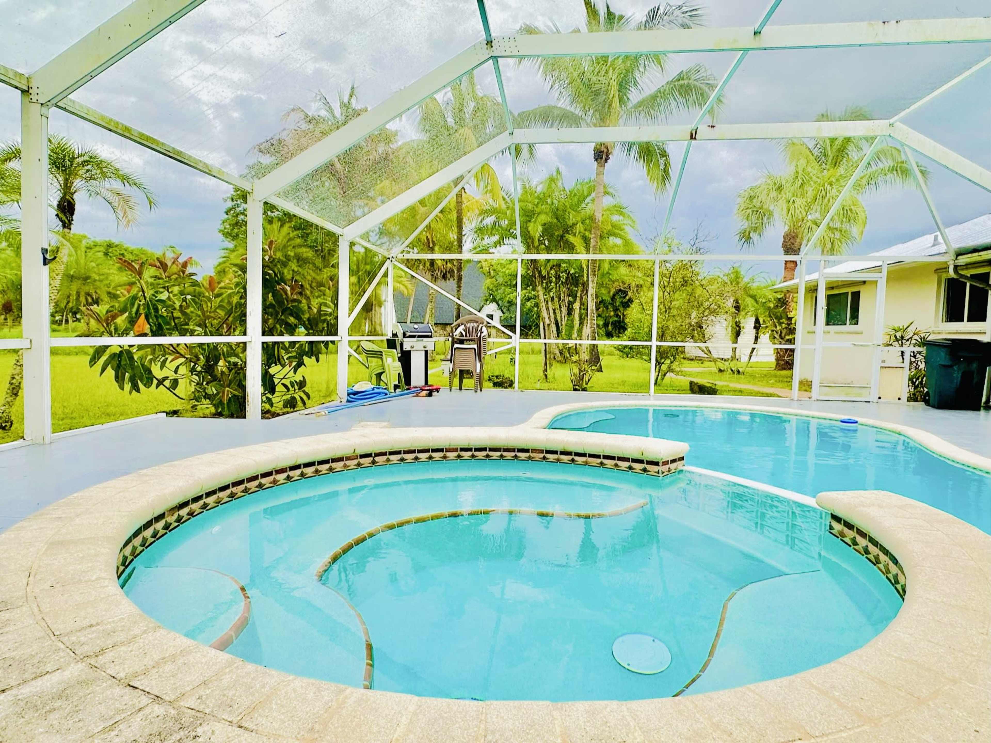 A screened-in pool area with a hot tub integrated into the pool design, surrounded by lush greenery and palm trees.