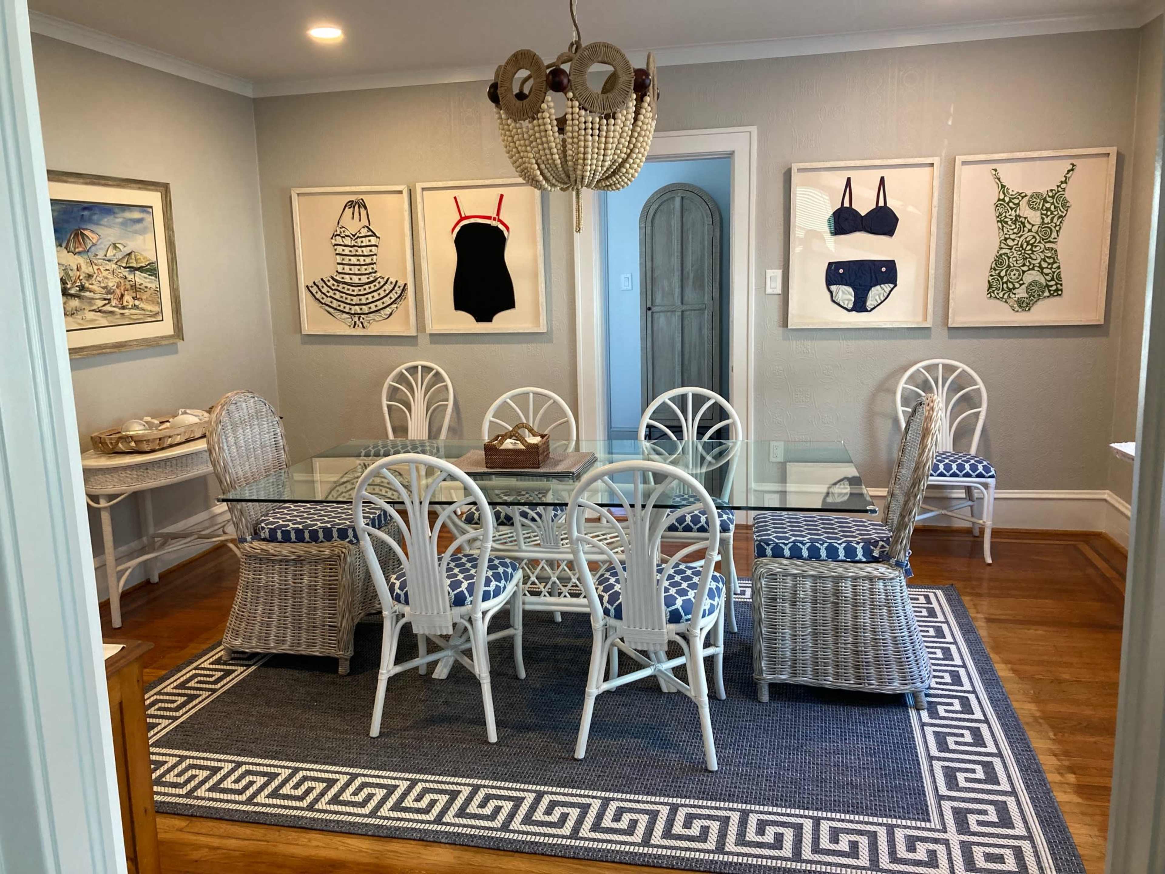 A dining room features a glass table surrounded by six white wicker chairs and two additional wicker chairs, with framed swimsuit artworks on the walls and a patterned rug underneath.