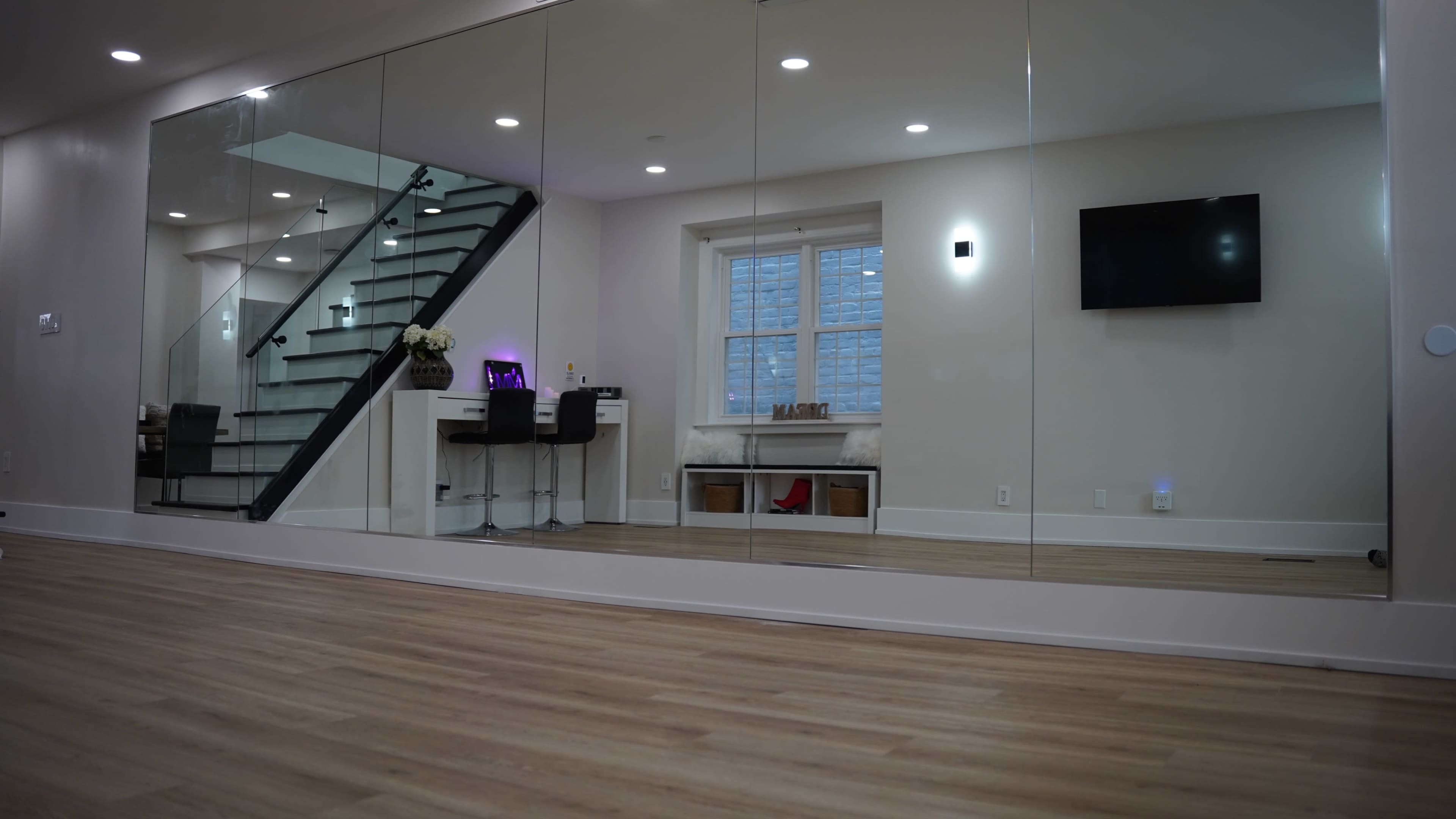 The image shows a modern room with wooden flooring, a large mirror on one wall, a staircase in the background, a workspace with two chairs, and a television mounted on another wall.