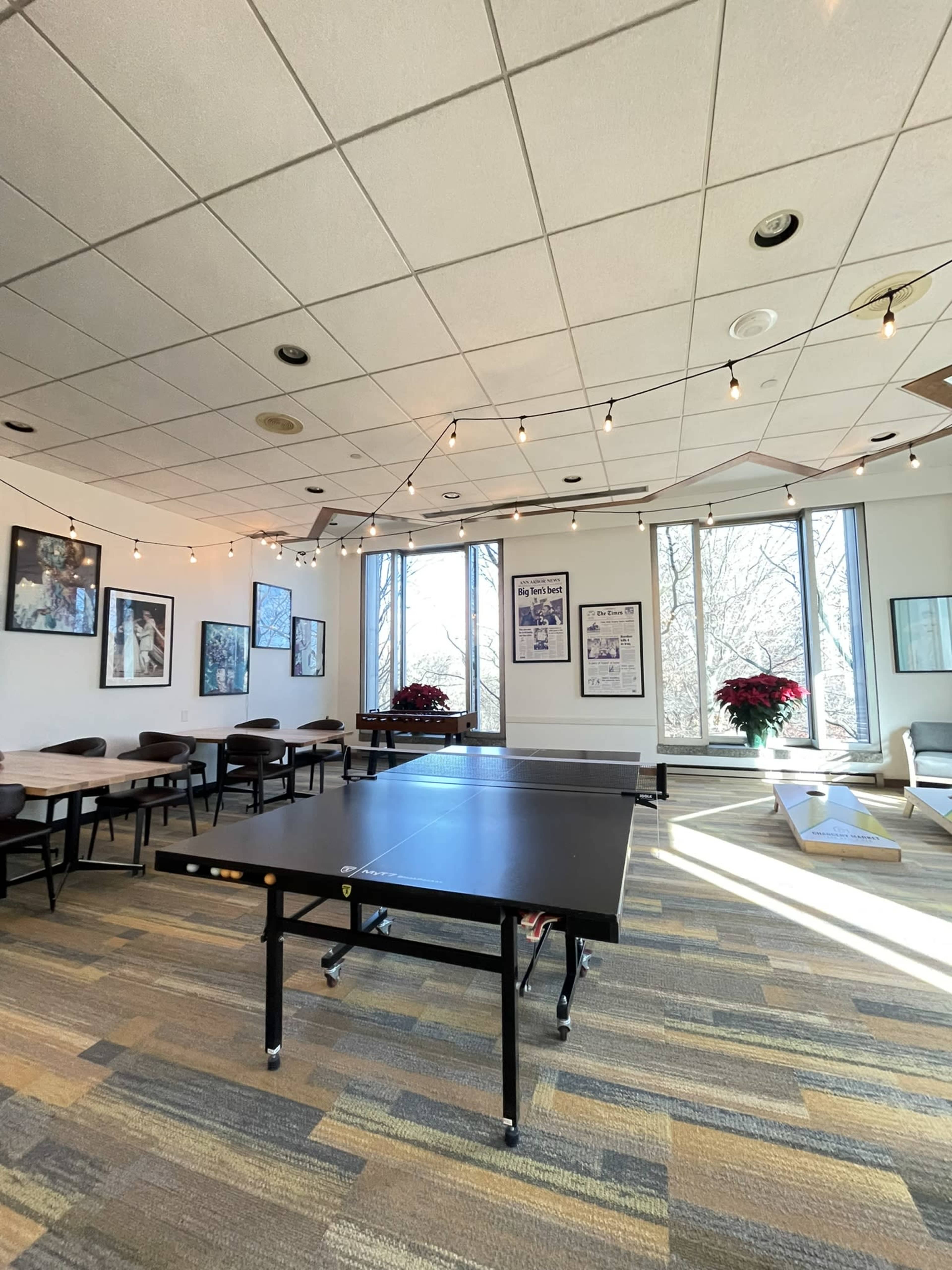 A spacious room features a ping pong table in the center, surrounded by tables and chairs, with large windows allowing natural light to illuminate the space.