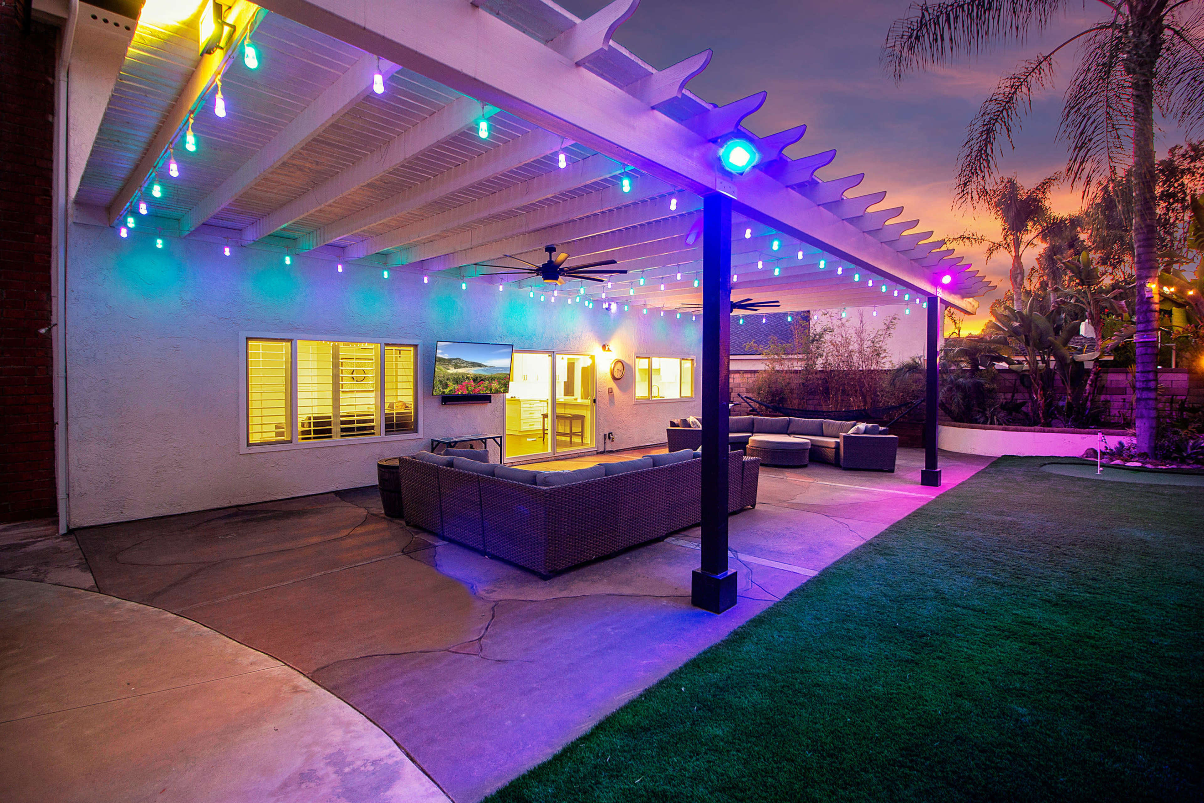 A patio area with a seated space, a television mounted on the wall, and colorful string lights illuminating the outdoor setting.