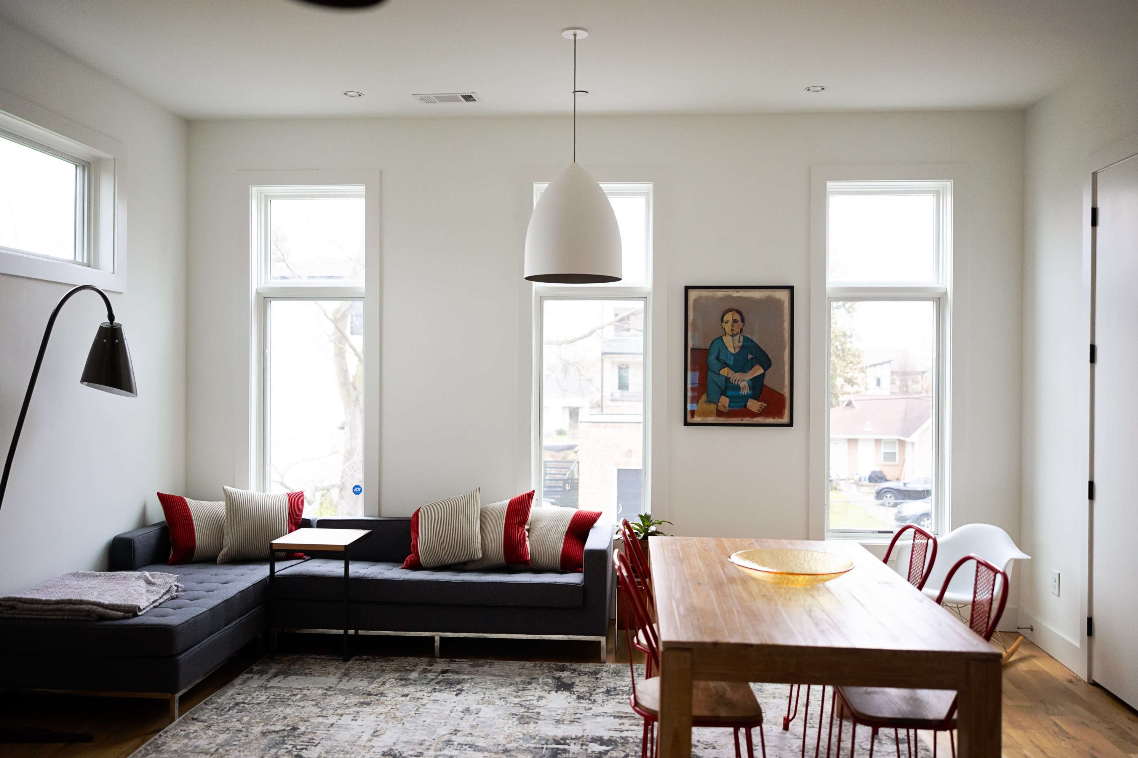 The image shows a modern living room with a sectional sofa, a wooden dining table surrounded by red chairs, and a framed artwork on the wall.