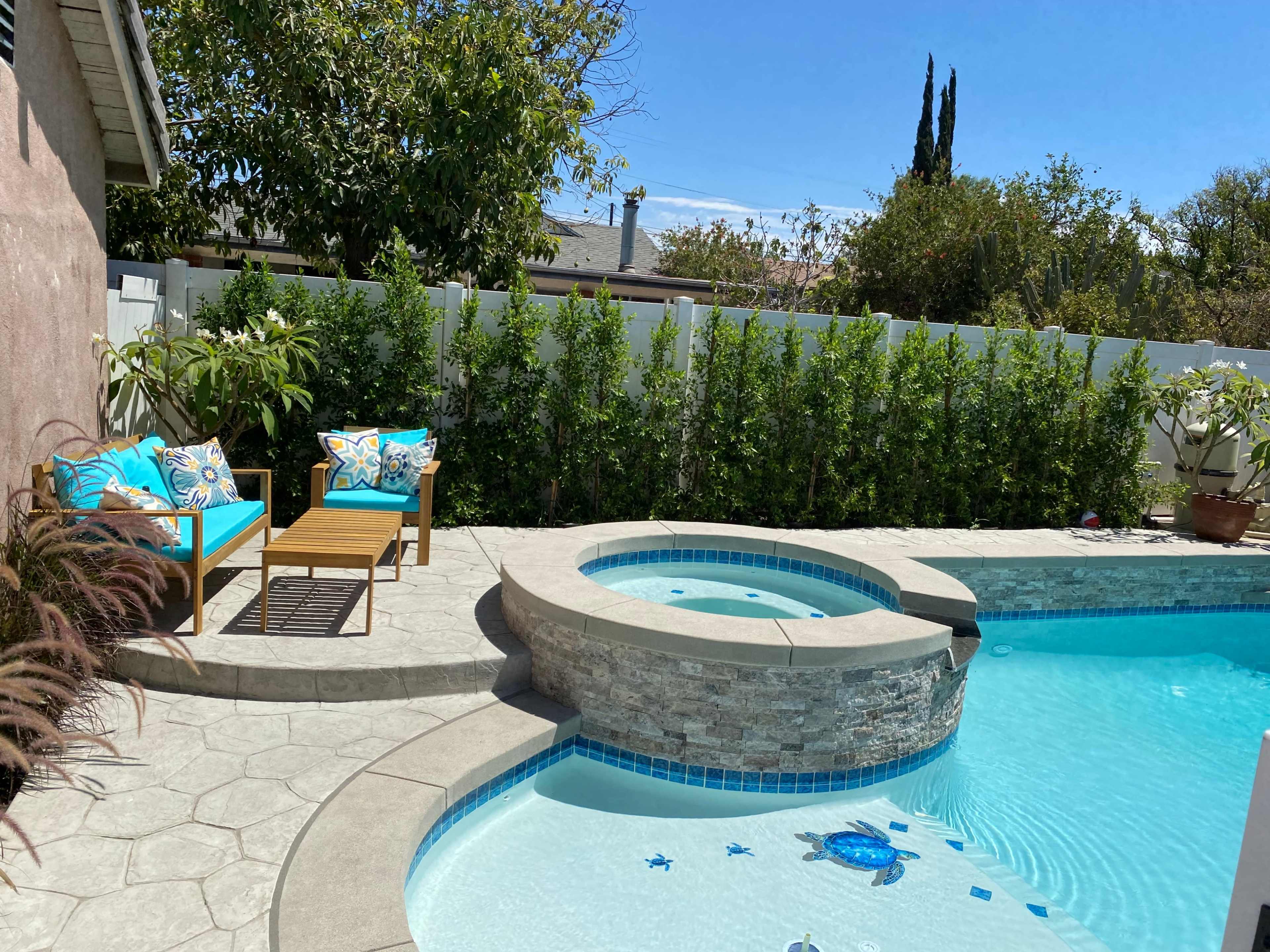 The scene shows a backyard pool area featuring a spa, decorative tiles, and two chairs with bright cushions beside a stone-lined pool.