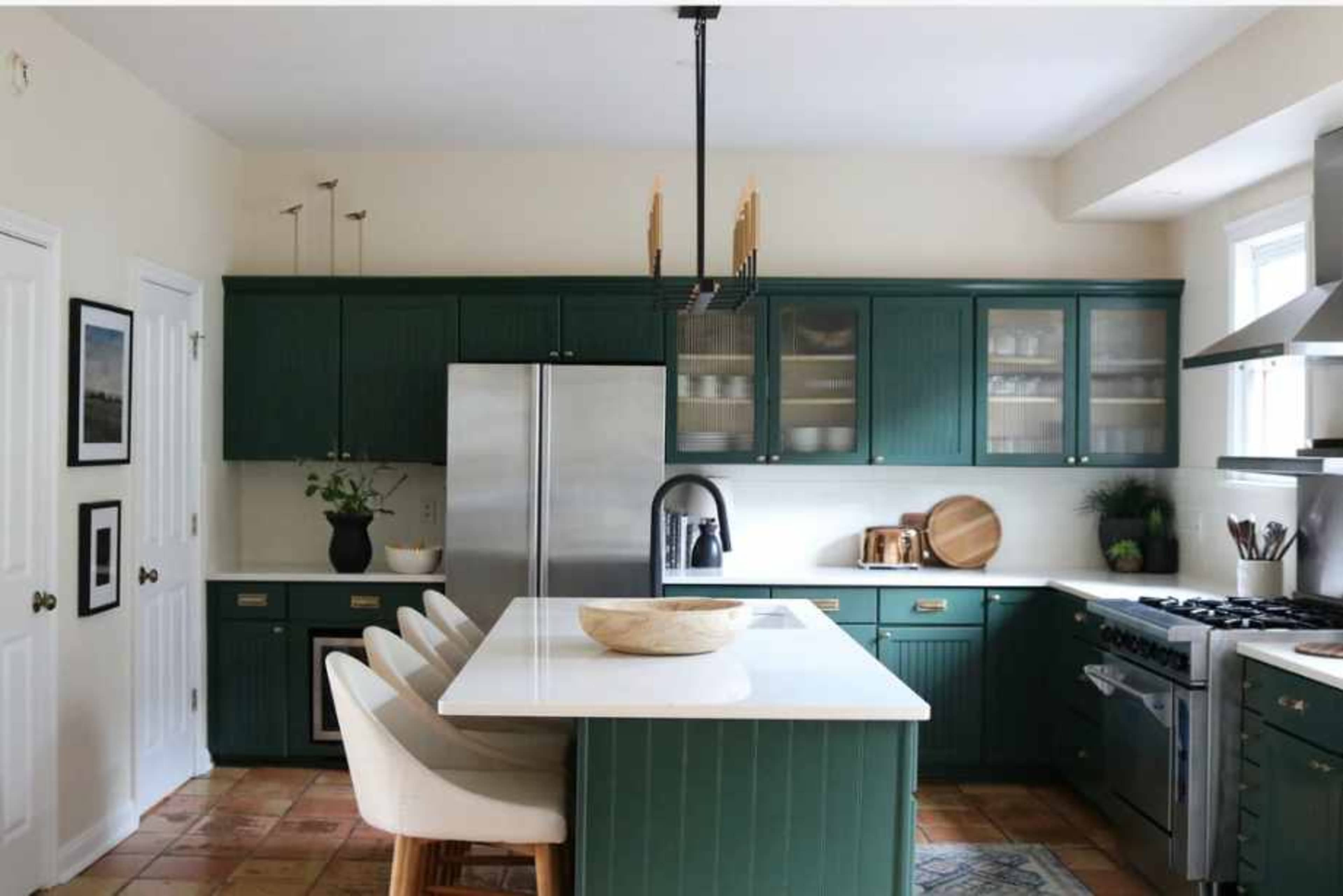 A modern kitchen features green cabinetry, a central island with seating, stainless steel appliances, and natural light streaming in from a window.