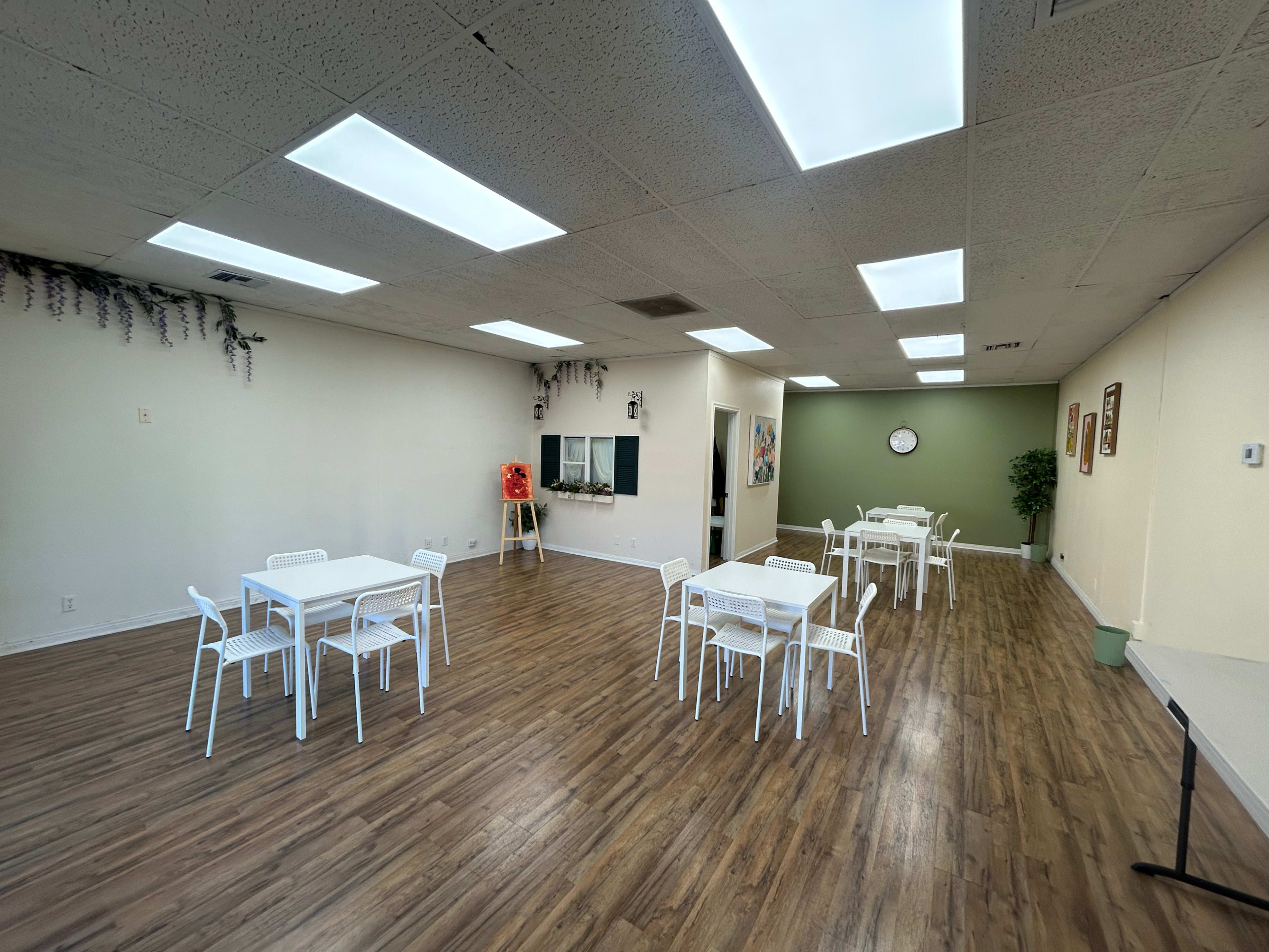 The image shows a spacious, well-lit room furnished with several white tables and chairs arranged in a simple layout.