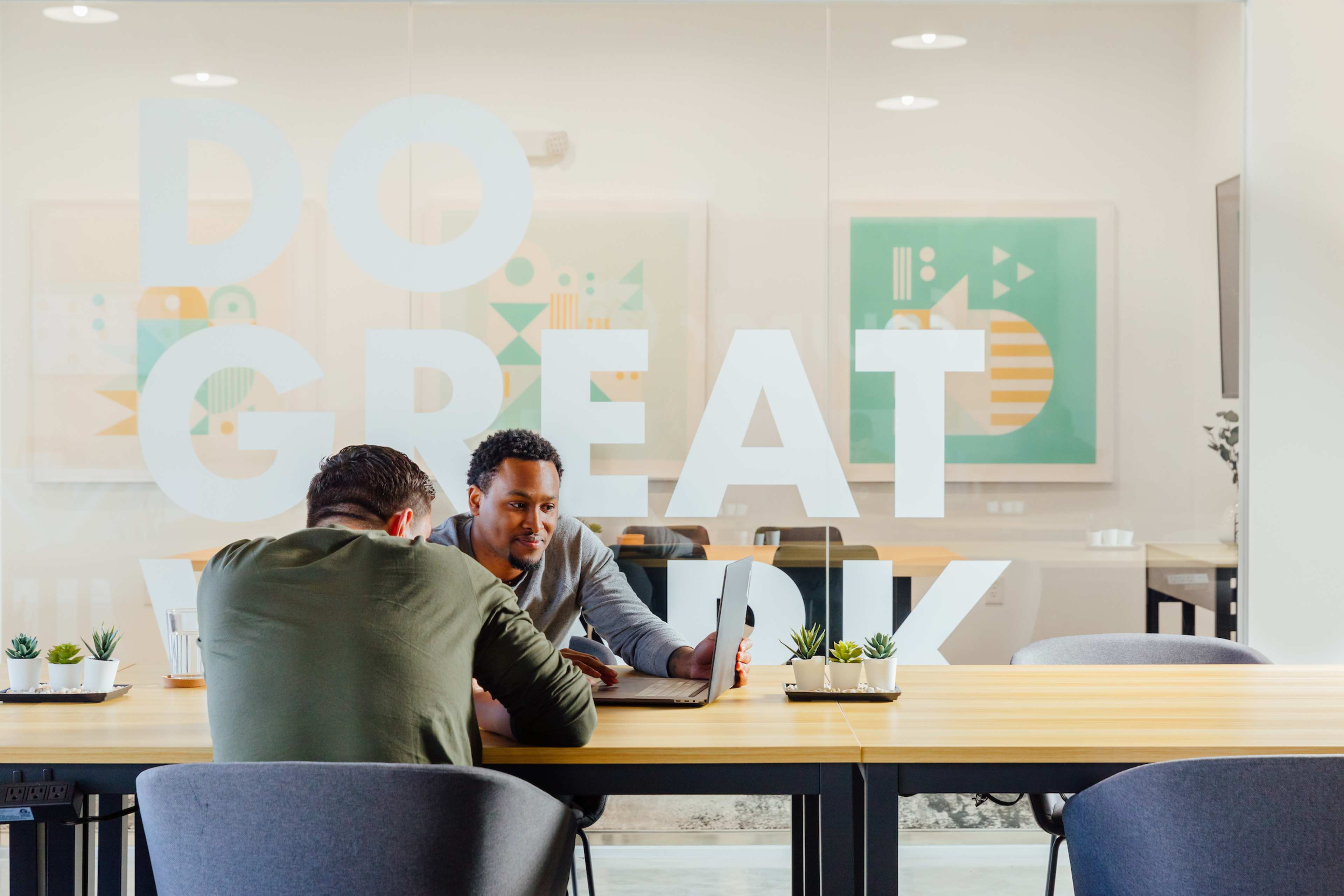 Two men are seated at a long table in a modern office space, collaborating on a laptop with large motivational phrases on a glass partition behind them.
