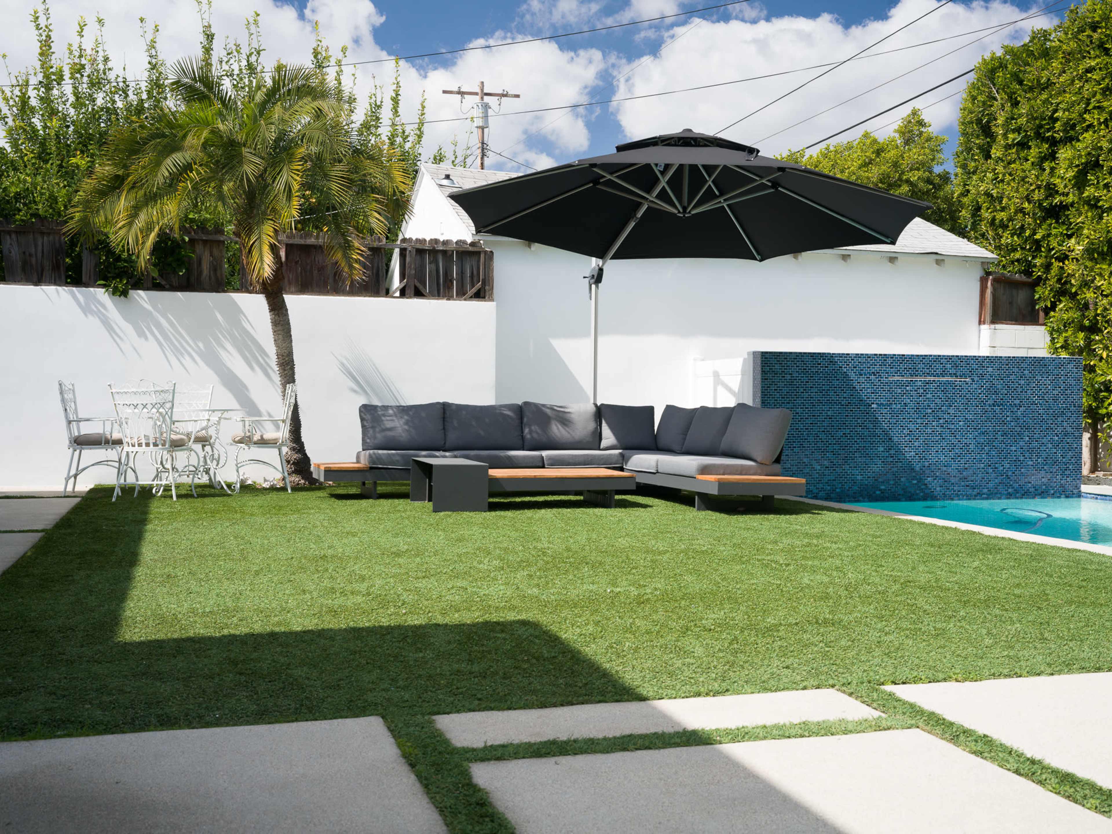 A patio area with a swimming pool, featuring a dark umbrella over a gray sofa, surrounded by green grass and a white wall.