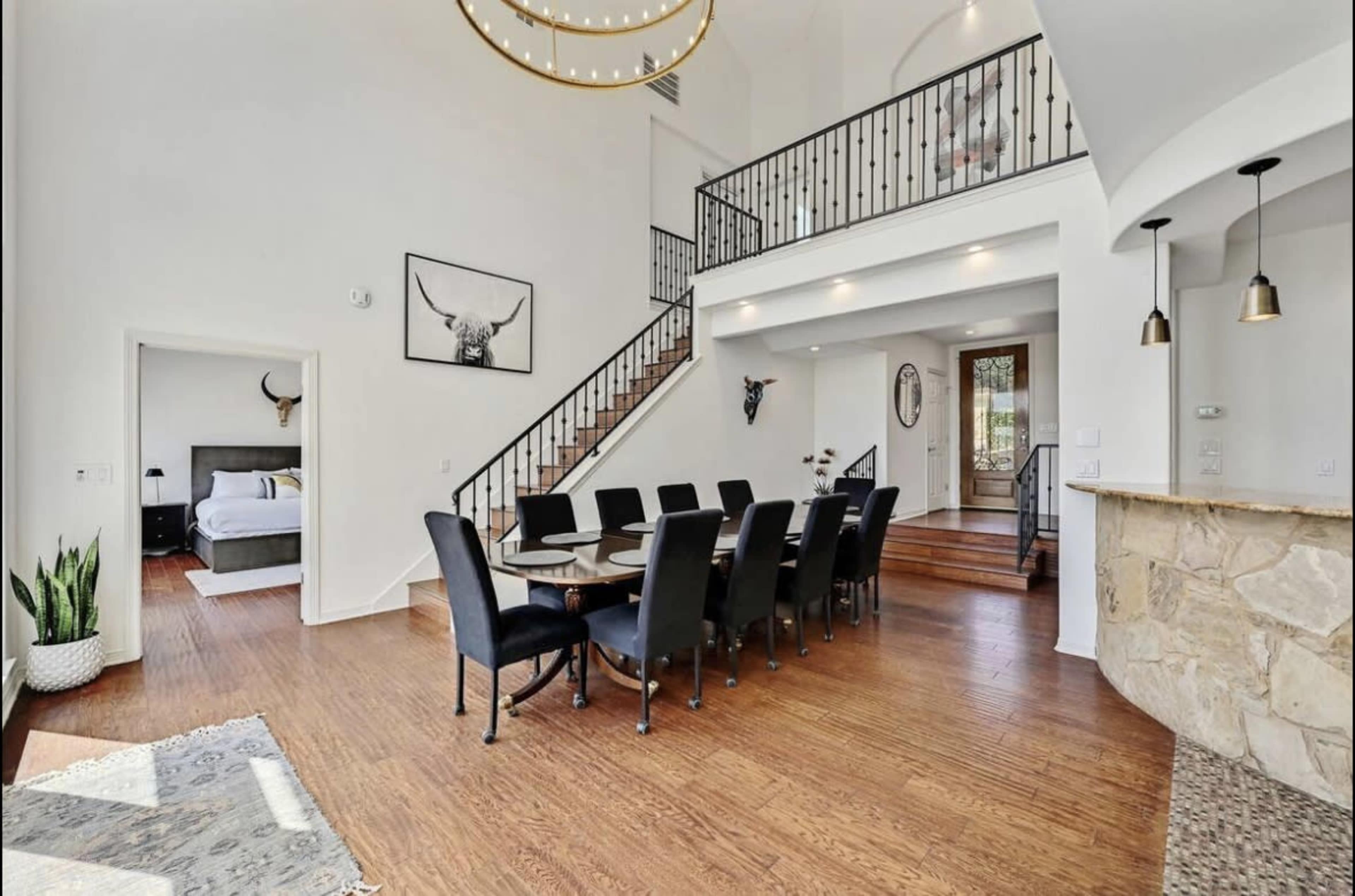 A spacious dining area with a large table and black chairs is positioned next to a staircase leading to an upper level, all set in a well-lit room with high ceilings.